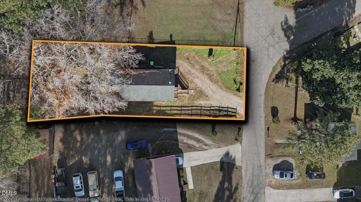 Aerial view of a rectangular property outlined in orange, containing a house, a driveway, and trees. The property is bordered by streets on two sides, with parked cars and other houses nearby.