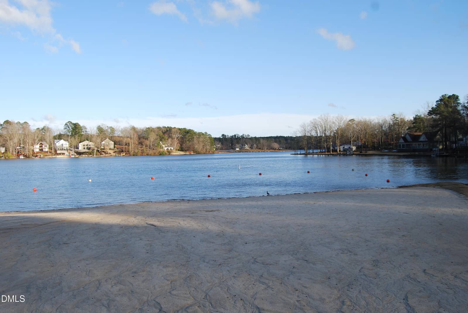 A sandy beach meets a calm lake bordered by floating red buoys. Houses and trees line the opposite shore under a mostly clear blue sky with some clouds.