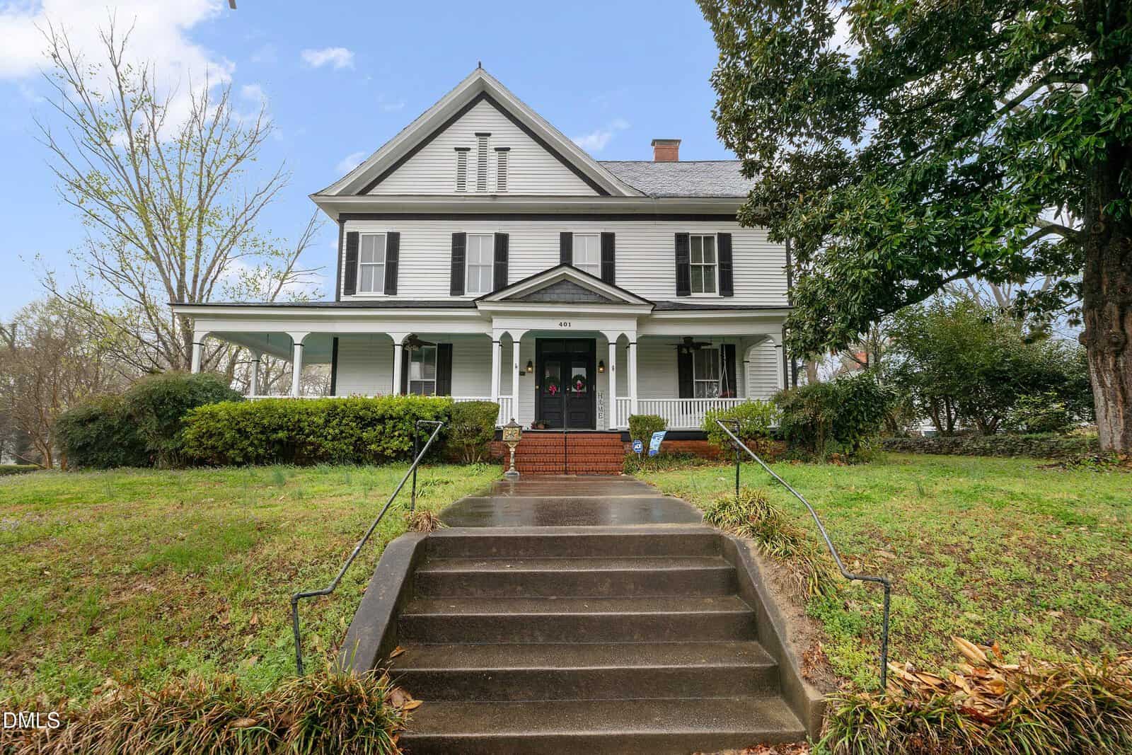 A two-story white Victorian house with black shutters, a covered front porch, and brick steps leading up from a sidewalk on a grassy lawn, surrounded by trees and shrubs.