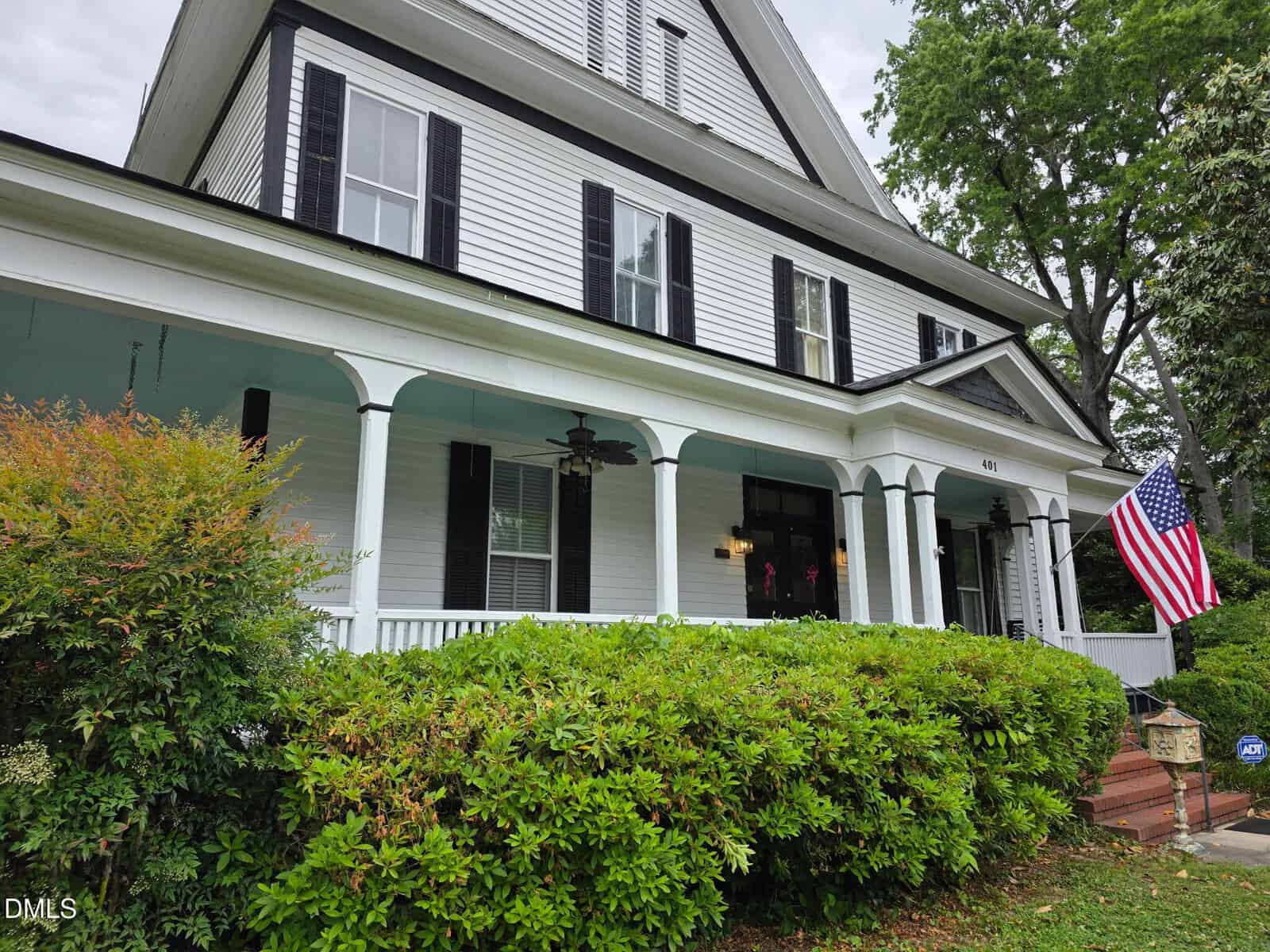 White two-story house with black shutters, a covered porch, ceiling fans, and an American flag. Lush green bushes and trees surround the front steps. House number 401 is visible above the porch.