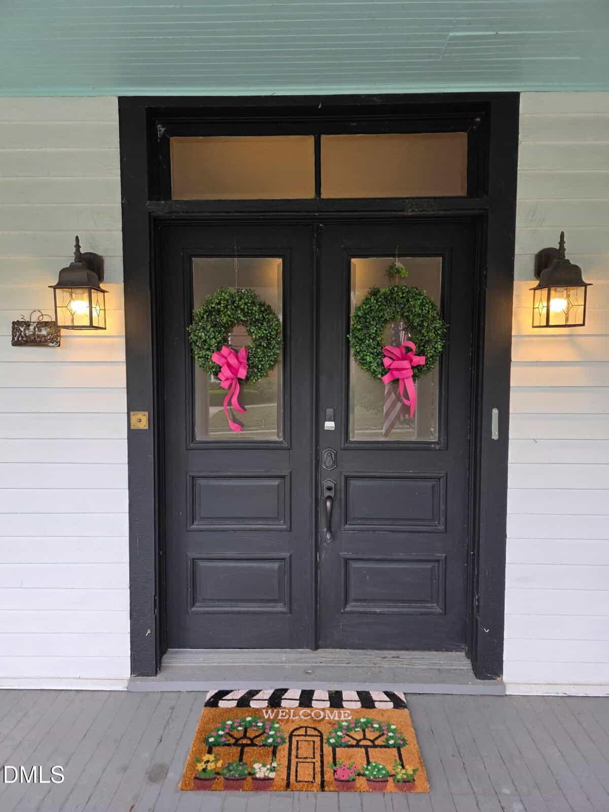 A pair of black double front doors with green wreaths and pink bows, flanked by wall lanterns. A colorful "Welcome" doormat with potted plants is on the gray porch floor. The house exterior is painted white.