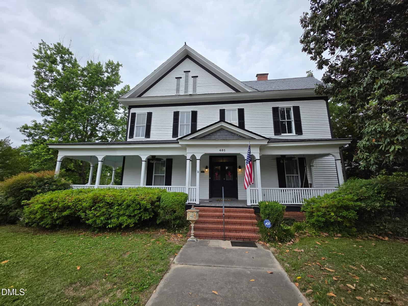 A large white two-story house with black shutters, a front porch, an American flag, and steps leading to the entrance. The yard has green grass, bushes, and trees, under a cloudy sky.