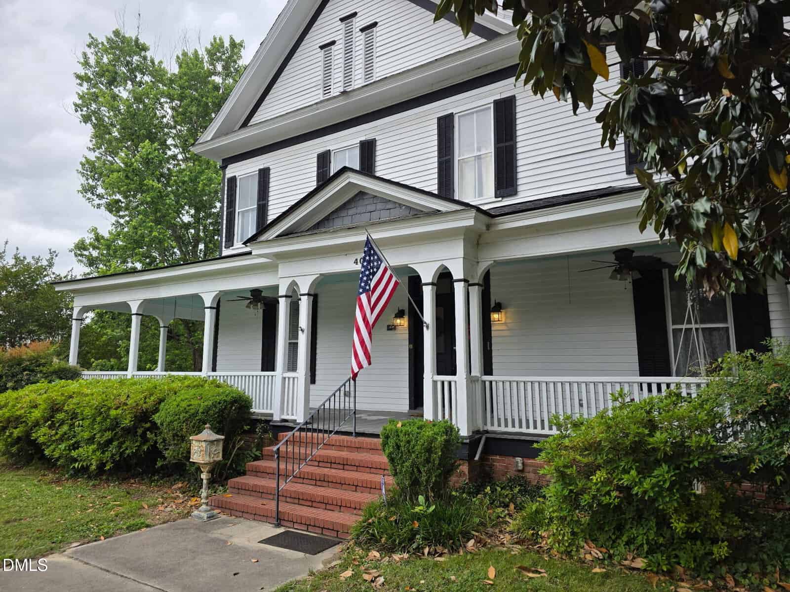 A white, two-story house with black shutters and a covered front porch sits at 401 N Main Street in Louisburg. An American flag hangs near the steps, bordered by green bushes. The yard features trees and a sidewalk.
