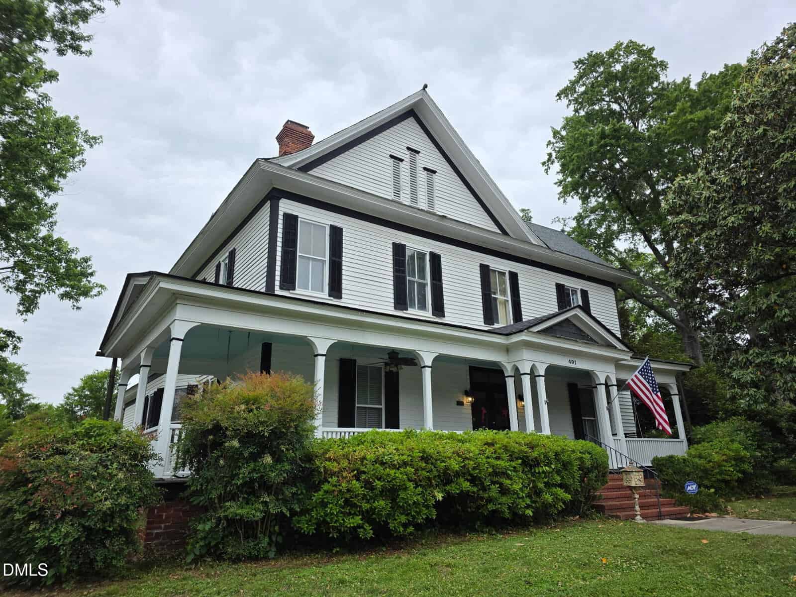 A large white two-story house with black shutters, a wraparound porch, and an American flag, surrounded by green bushes and trees under a cloudy sky.
