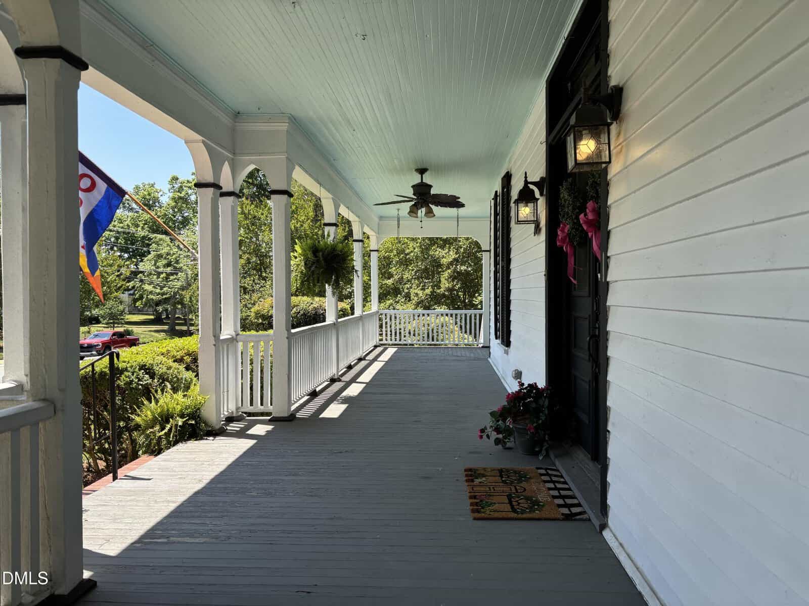 A spacious, covered front porch with white railings, black door, hanging flower baskets, ceiling fans, and a colorful flag. Sunlight casts shadows, and greenery is visible in the yard beyond.