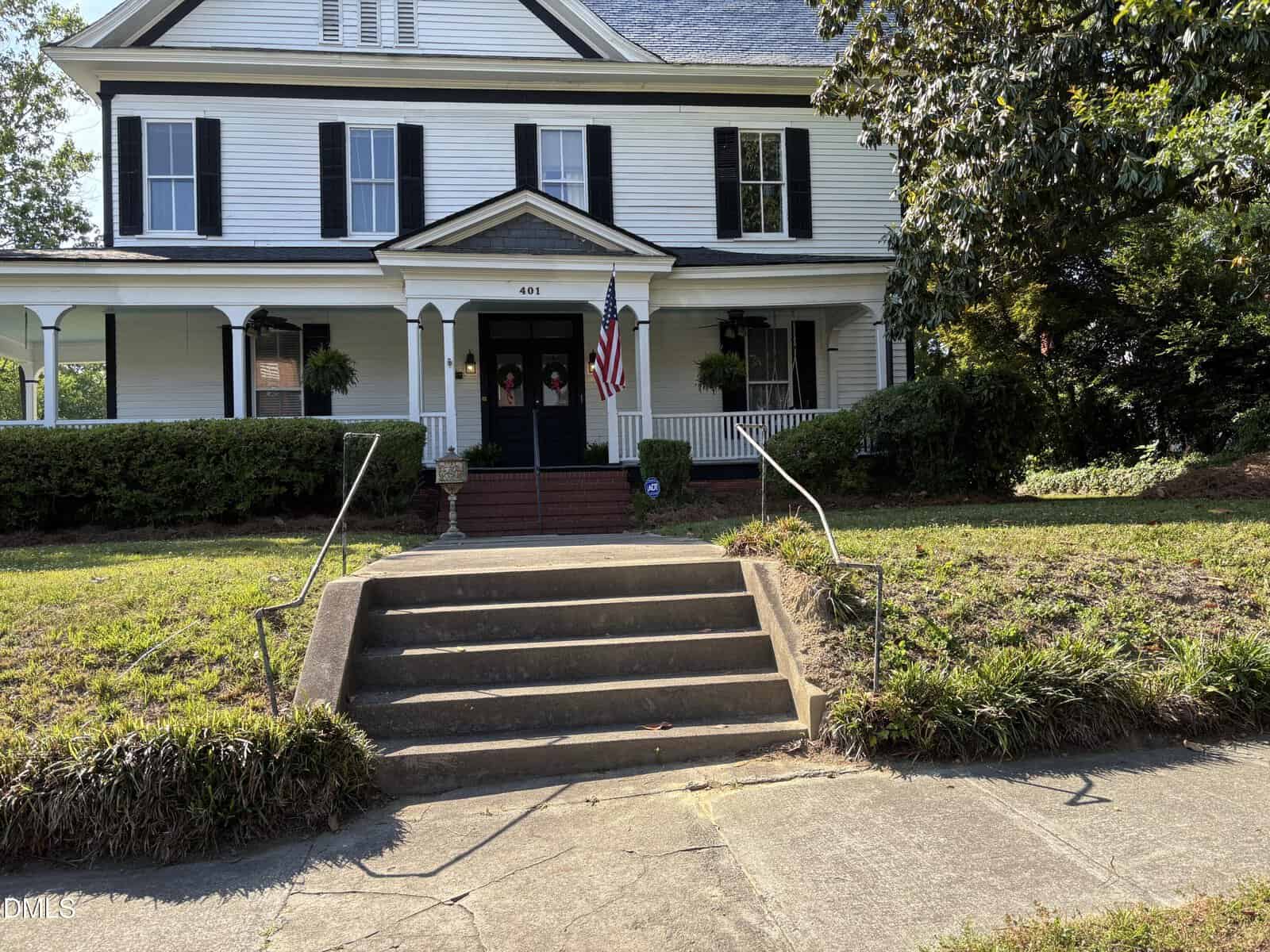 A large white house with black shutters, a front porch, and an American flag. A concrete sidewalk and steps lead up to the porch, with green lawns and bushes on either side.