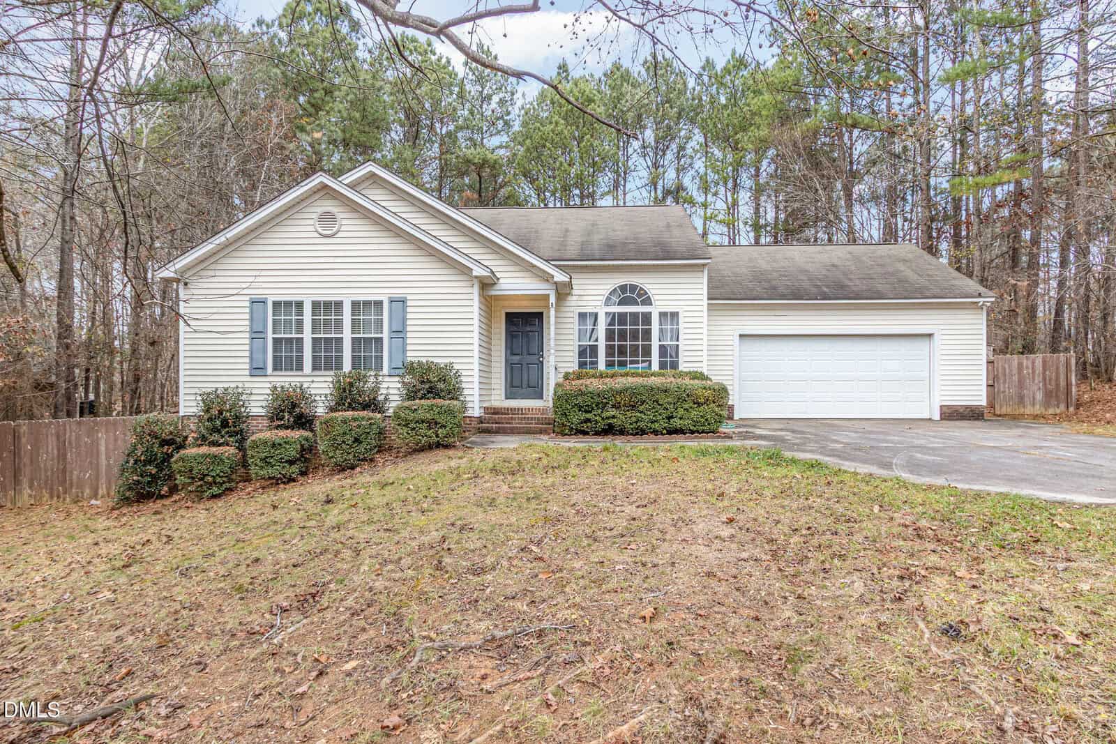 Single-story house at 60 Coral Ridge Circle, Franklinton, with light-colored siding, blue shutters, and a dark front door. There is a two-car garage, shrubbery in front, a concrete driveway, and trees in the background. The yard has patchy grass and a gentle slope.