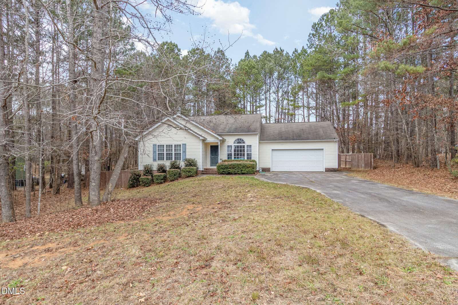 Single-story house with white siding and a two-car garage, set back from a paved driveway. The yard is mostly grass with some bare patches, and the house is surrounded by tall trees in a wooded area.