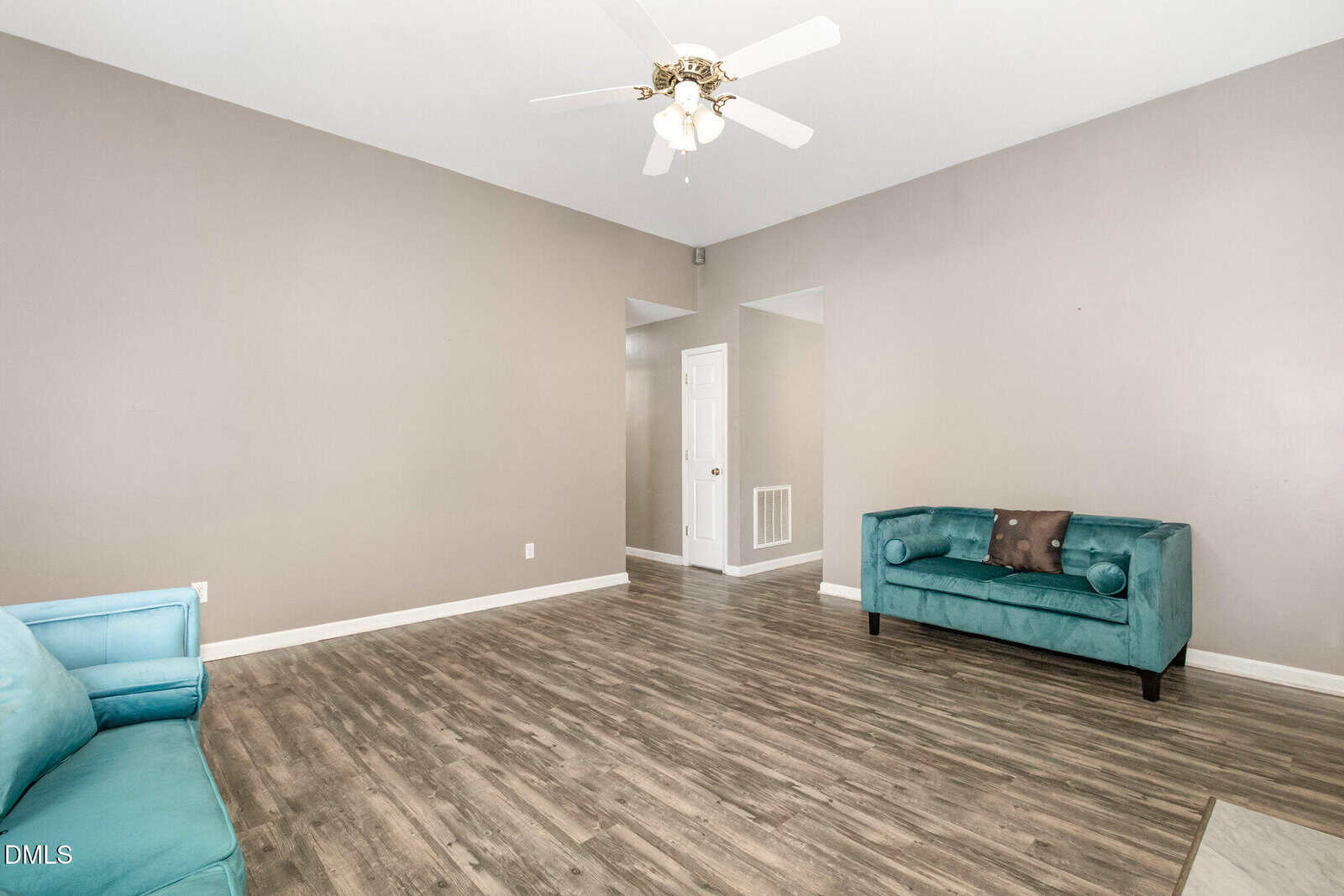 Living room with light brown walls, wood-style flooring, white ceiling fan with light, and two turquoise sofas. A white door and hallway are visible in the background. Room is sparsely furnished.