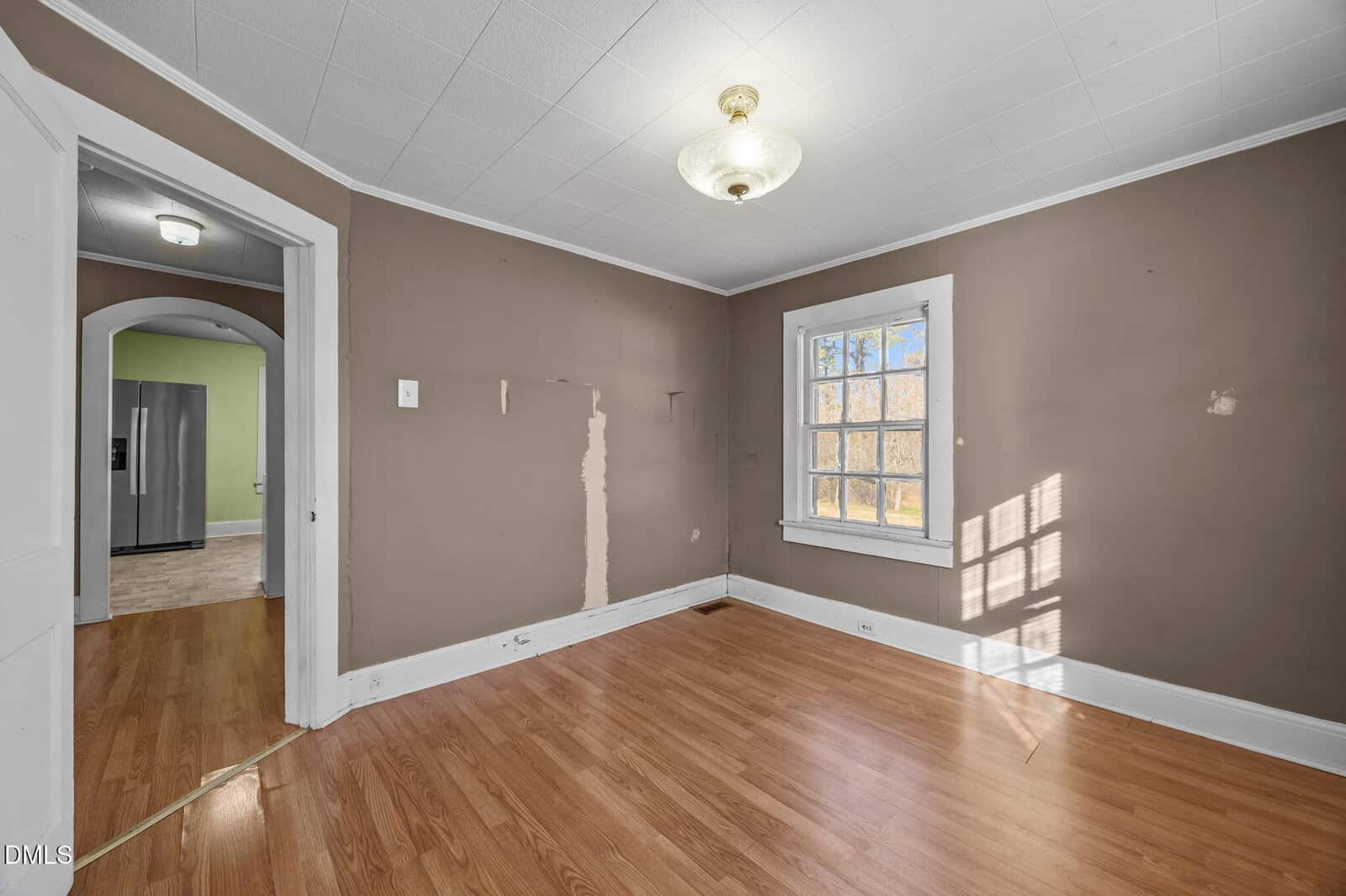 An empty room with brown walls, white trim, wood flooring, a single window, ceiling light, and an entryway leading to a kitchen with a refrigerator. There is a visible patch on one wall.
