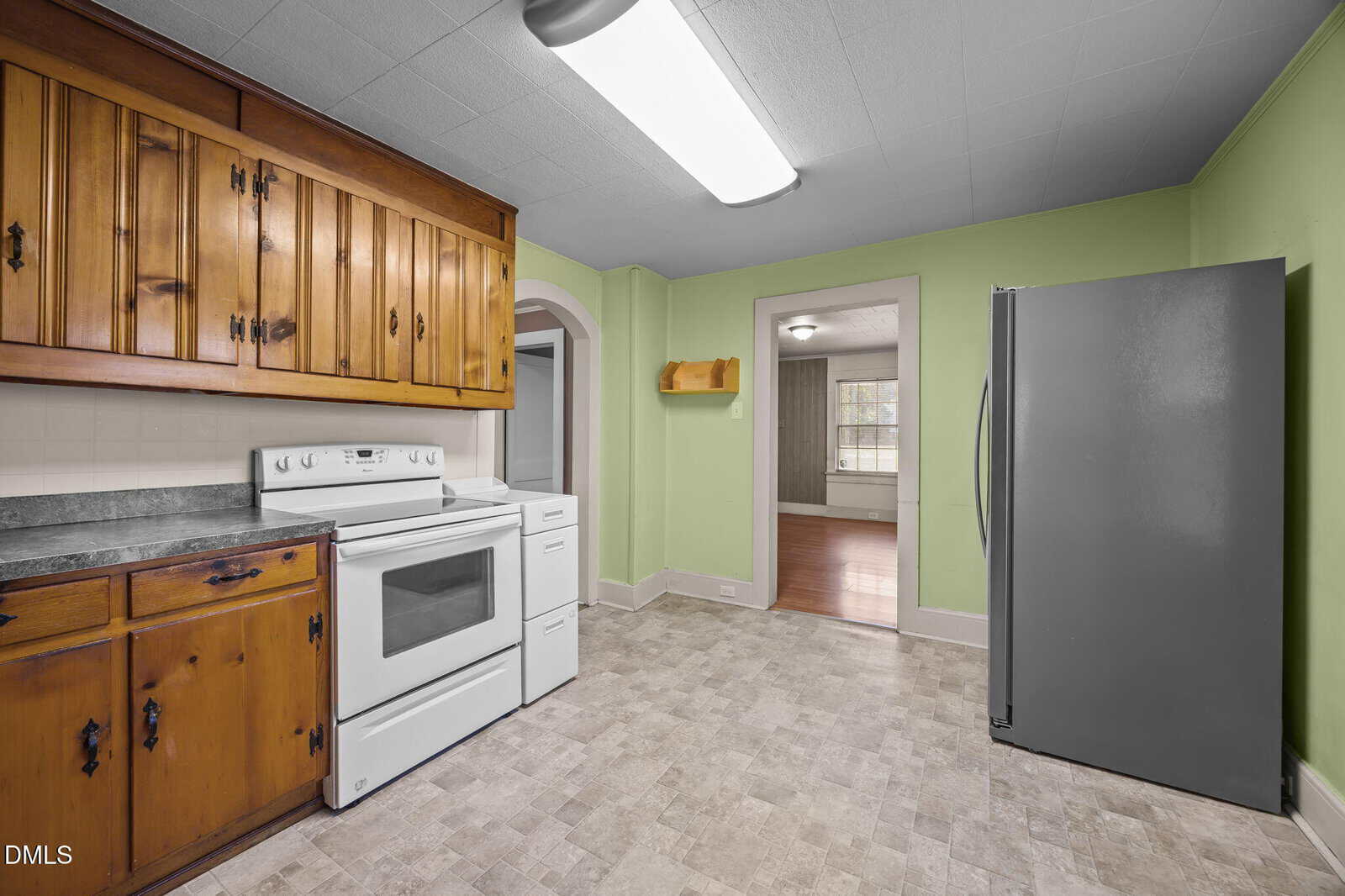 A kitchen with green walls, wooden cabinets, white electric stove, and refrigerator. The floor has a light-colored tile pattern. An arched doorway leads to an adjacent room with hardwood flooring and large window.