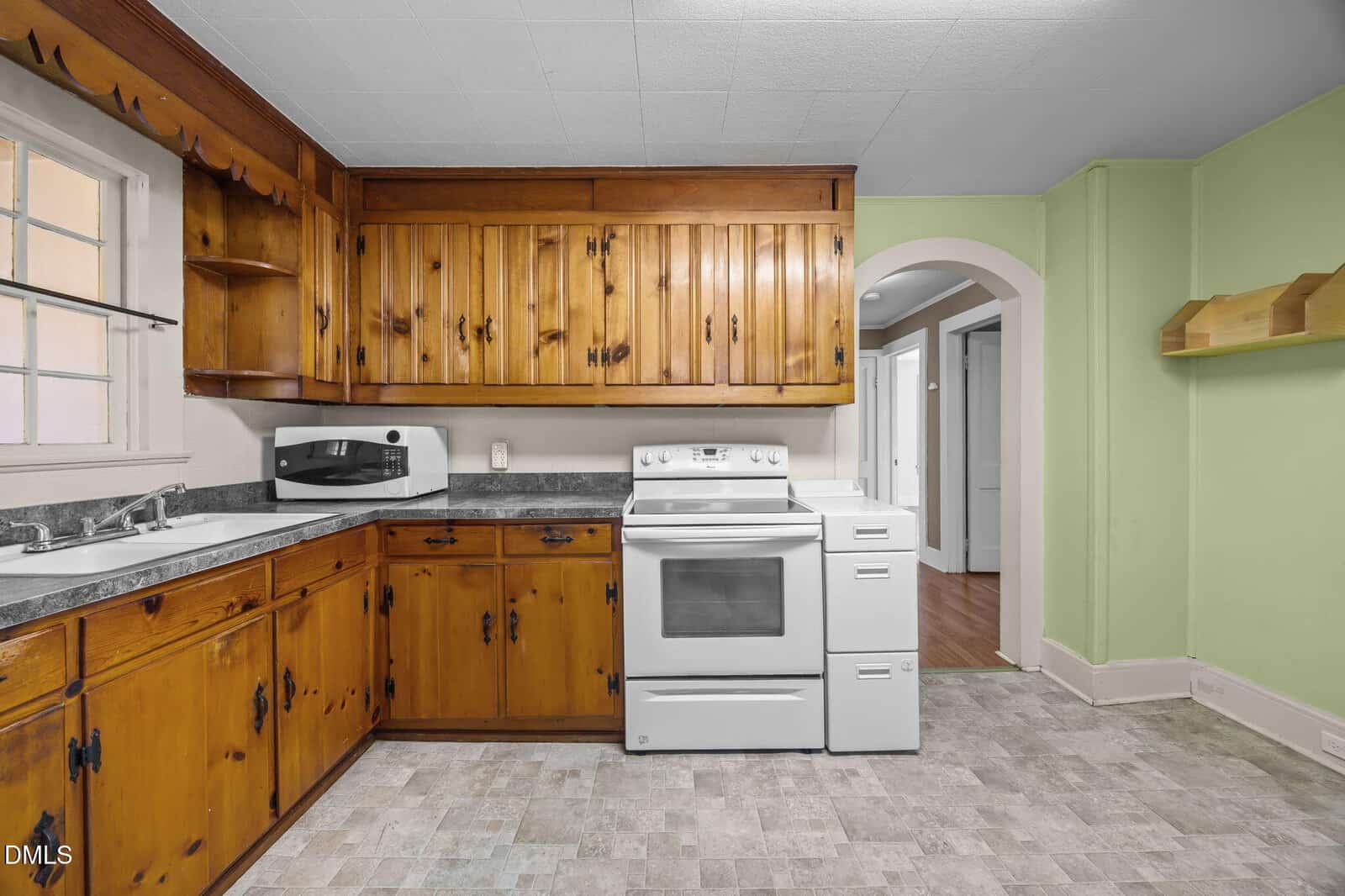 A kitchen with wood cabinets, white appliances including a stove and microwave, a double sink, green walls, and an arched doorway leading to another room.