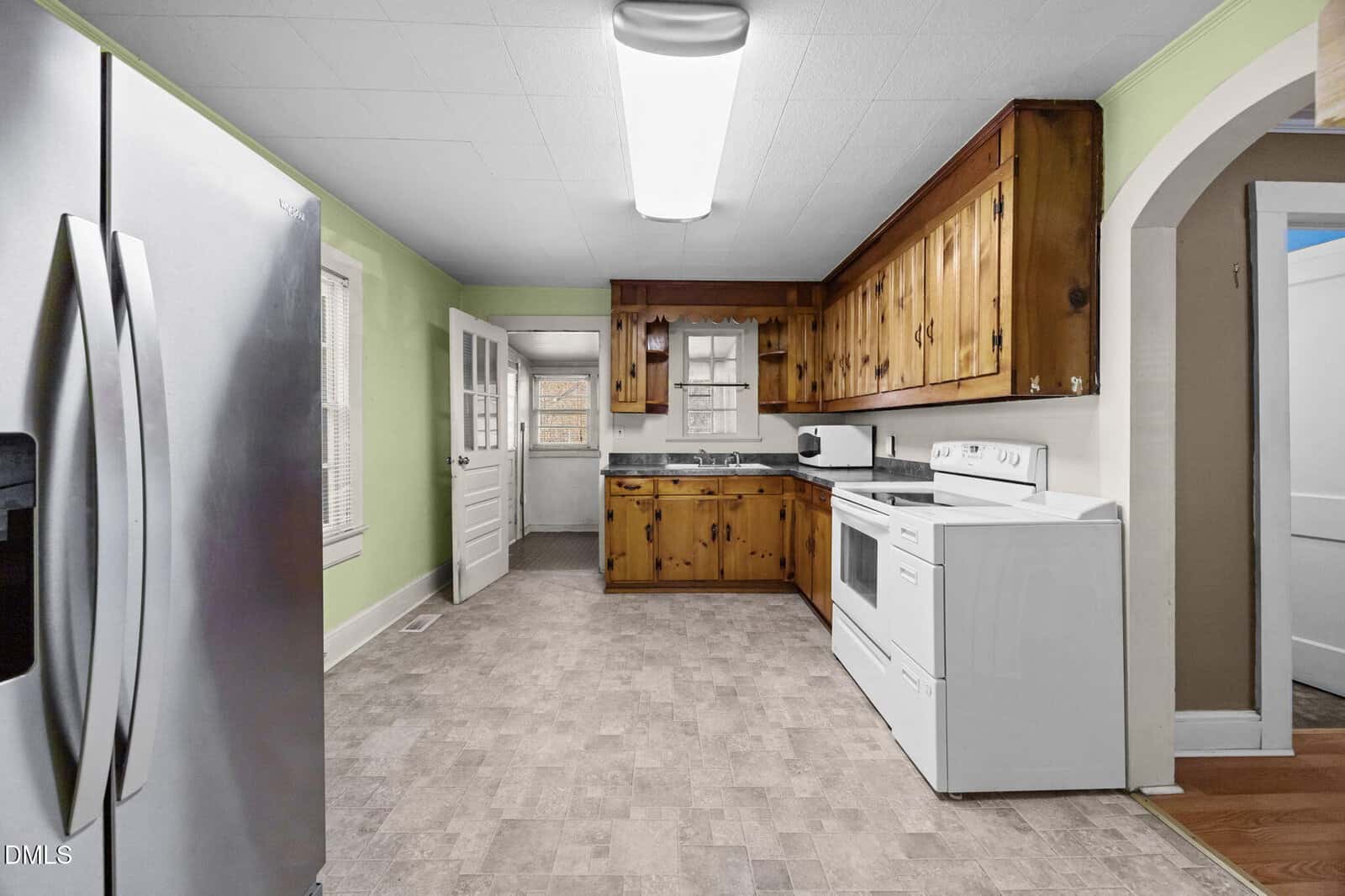 A kitchen with wood cabinets, white appliances including a stove and refrigerator, light green walls, a window, and a door leading outside. The floor has a gray patterned linoleum.