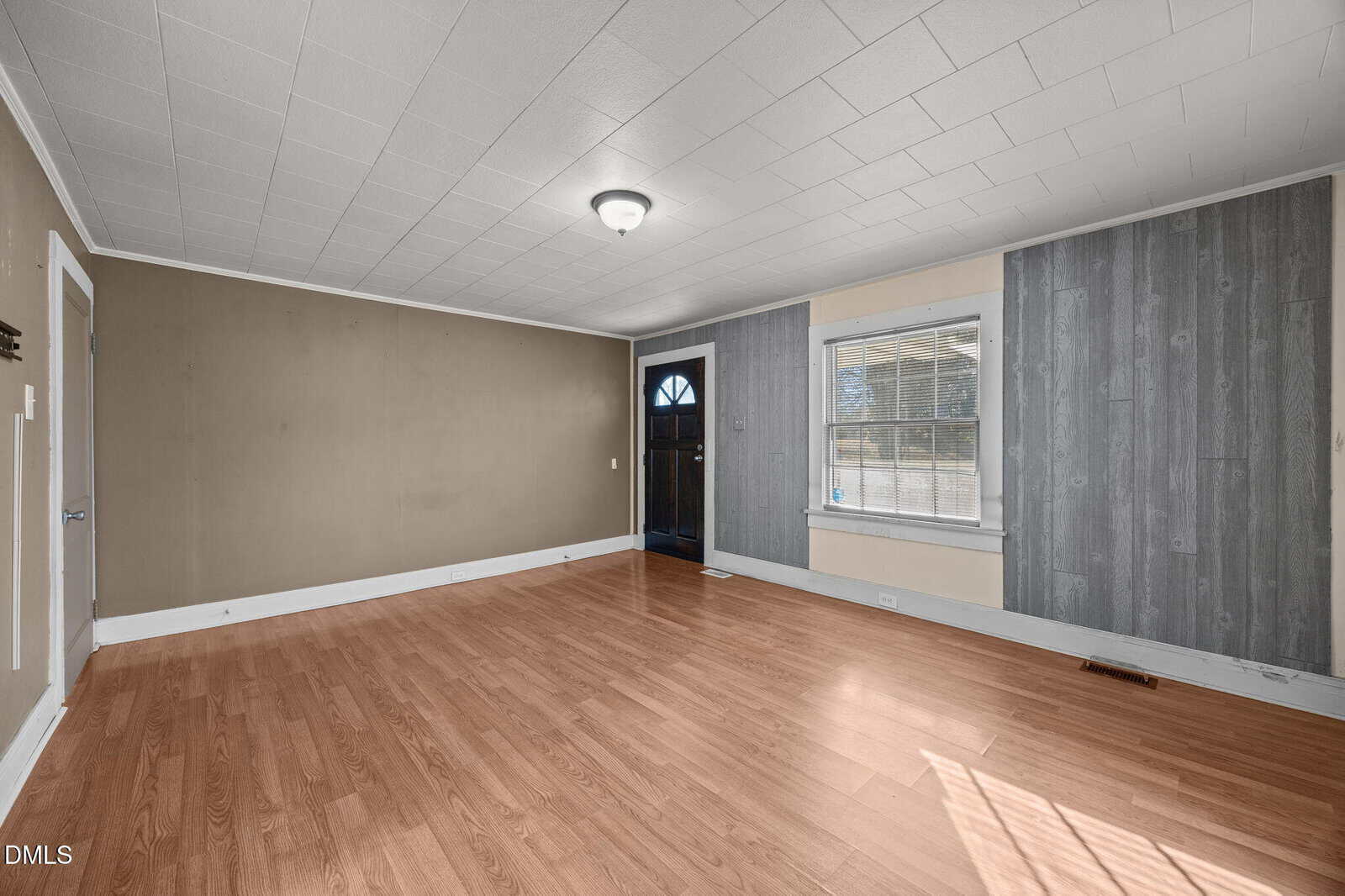 Empty room with light brown wood flooring, beige and gray paneled walls, white trim, a single ceiling light, a window with blinds, and a closed front door with a window insert.