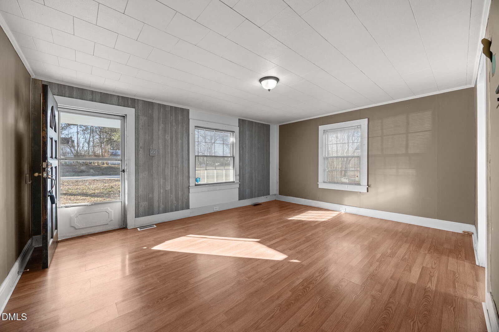 Empty living room with light brown laminate flooring, beige and gray walls, two large windows, a white ceiling with one light fixture, and a door with a window leading outside. Sunlight shines through the windows.