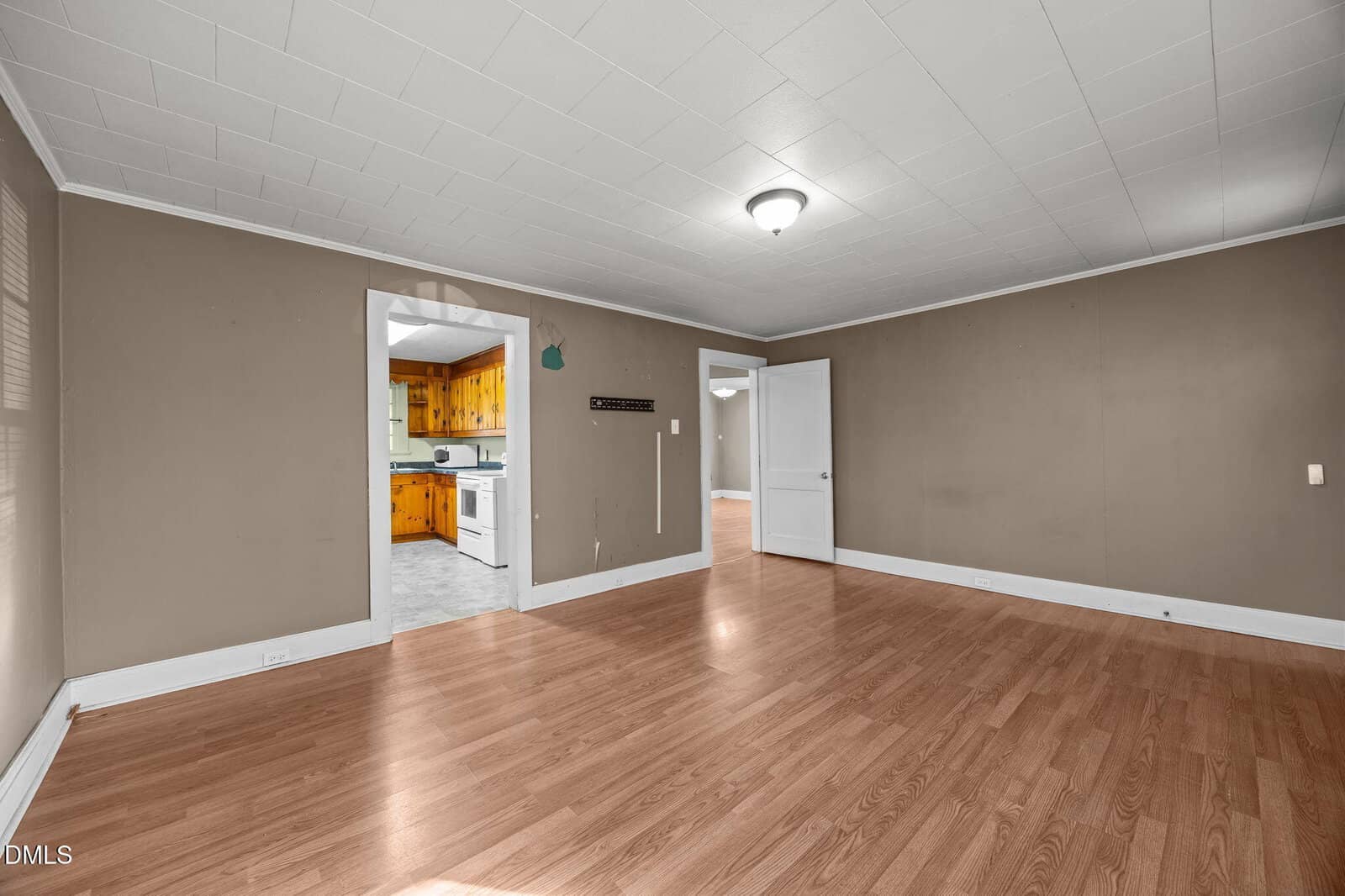 Empty room with wood laminate flooring, beige walls, white trim, and a white ceiling with a central light. Doorways lead to a kitchen with wood cabinets and another room with white walls and a window.