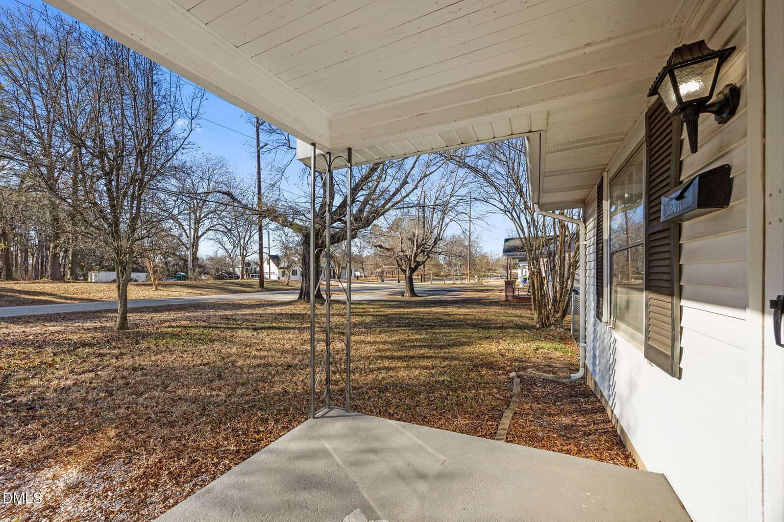 A covered front porch with a concrete floor, metal support posts, and an outdoor lantern attached to the white siding of a house. The yard has leaf-covered grass and several leafless trees. A street is visible in the background.