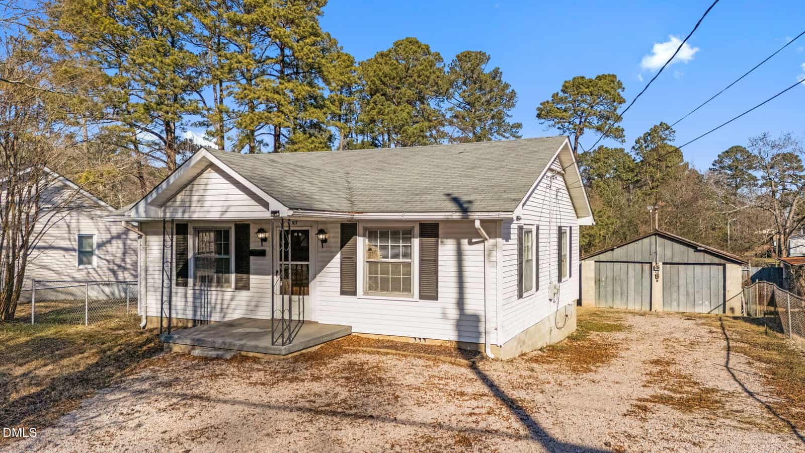 Single-story white house with gray roof, small covered front porch, and metal railing. Gravel driveway leads to a detached garage in the background. Pine trees and neighboring houses are visible.