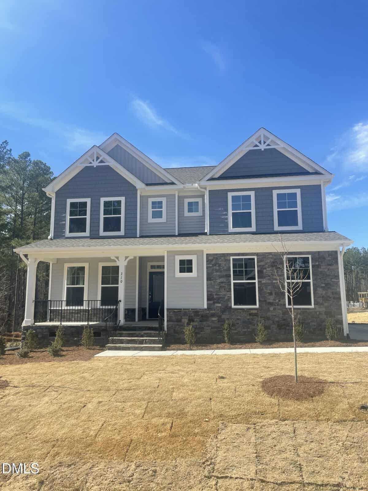Two-story house at 210 Ironwood Boulevard with gray siding and stone exterior accents, white trim, a covered front porch, multiple windows, and a bare lawn with new grass sod. Clear blue sky and trees are visible in the background of this beautiful Youngsville property.
