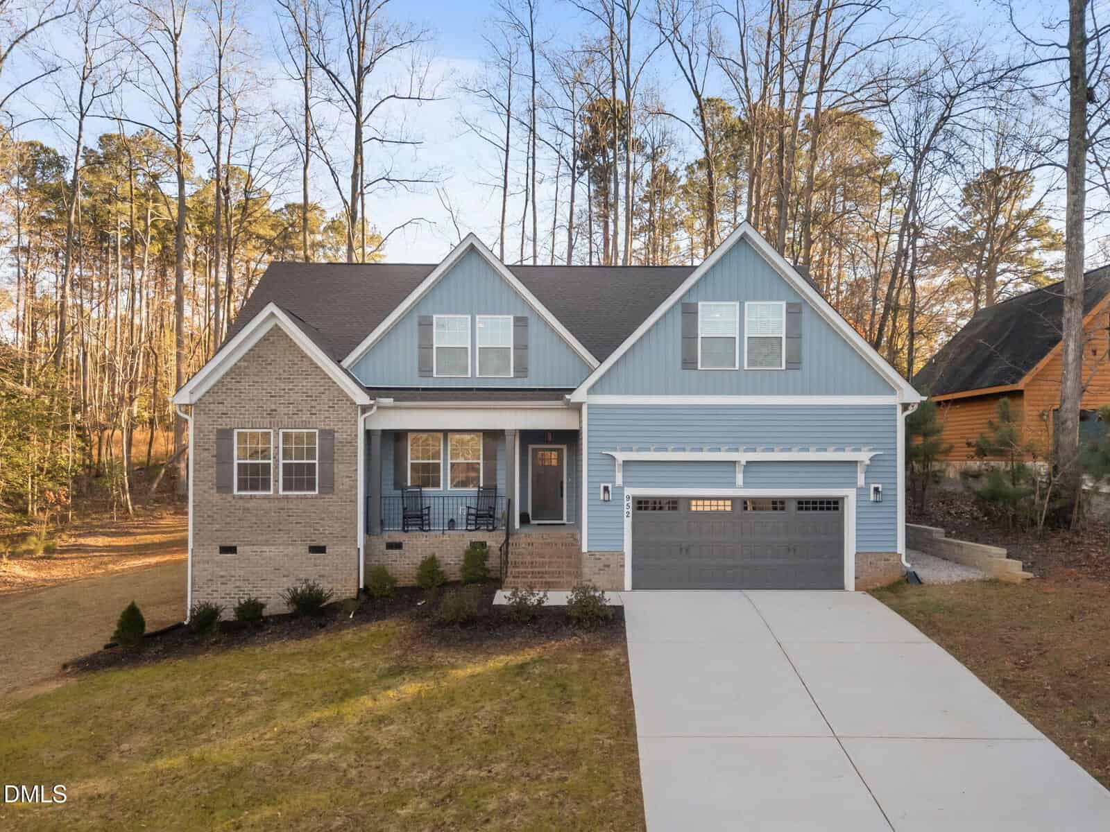 This two-story suburban house at 952 Sagamore Drive, Louisburg features blue siding, beige brick, a covered front porch, and a two-car garage. A sloped driveway leads up to the home, which is surrounded by trees and a spacious lawn.