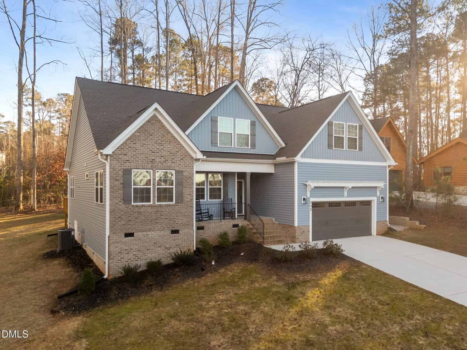 A two-story suburban house with a mix of brick and blue siding, a covered front porch, and a two-car garage. The house sits on a grassy lawn with trees in the background and a paved driveway in front.