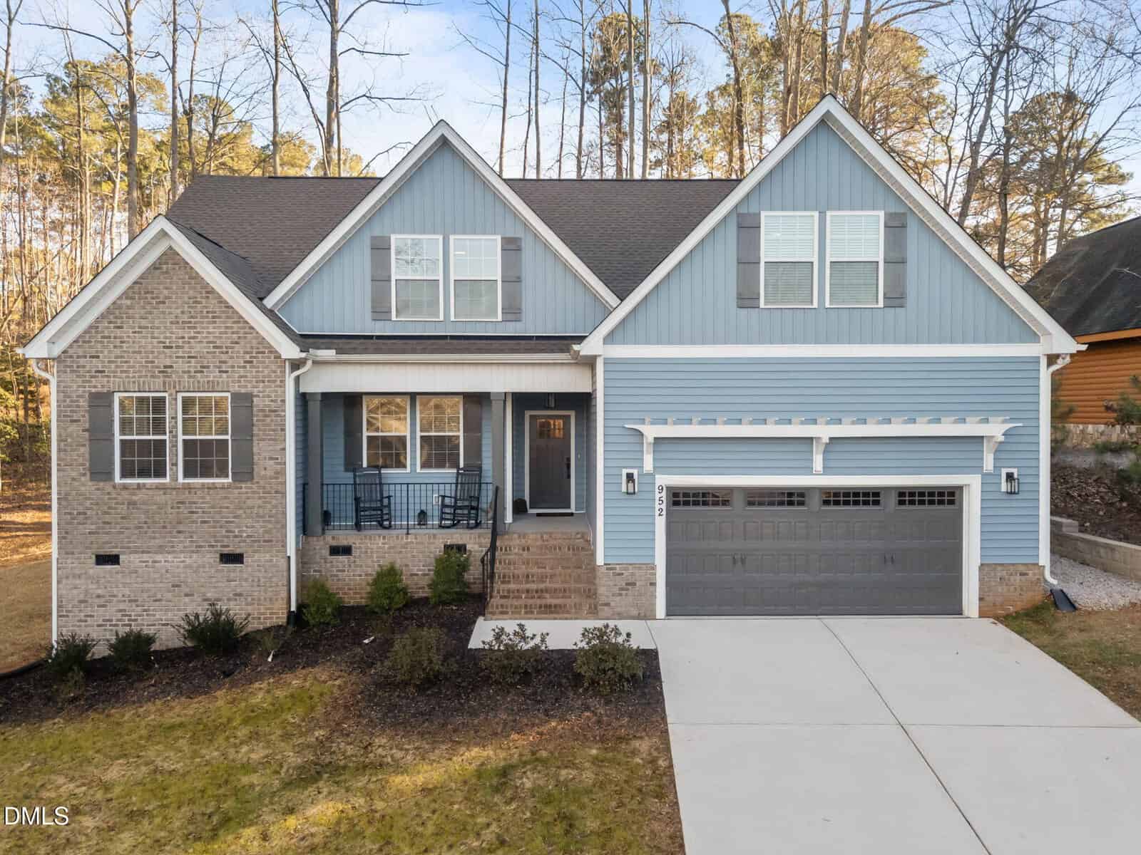 A two-story house with light brick and blue siding, a covered front porch with chairs, a dark gray double garage door, and a concrete driveway. Trees are visible in the background.