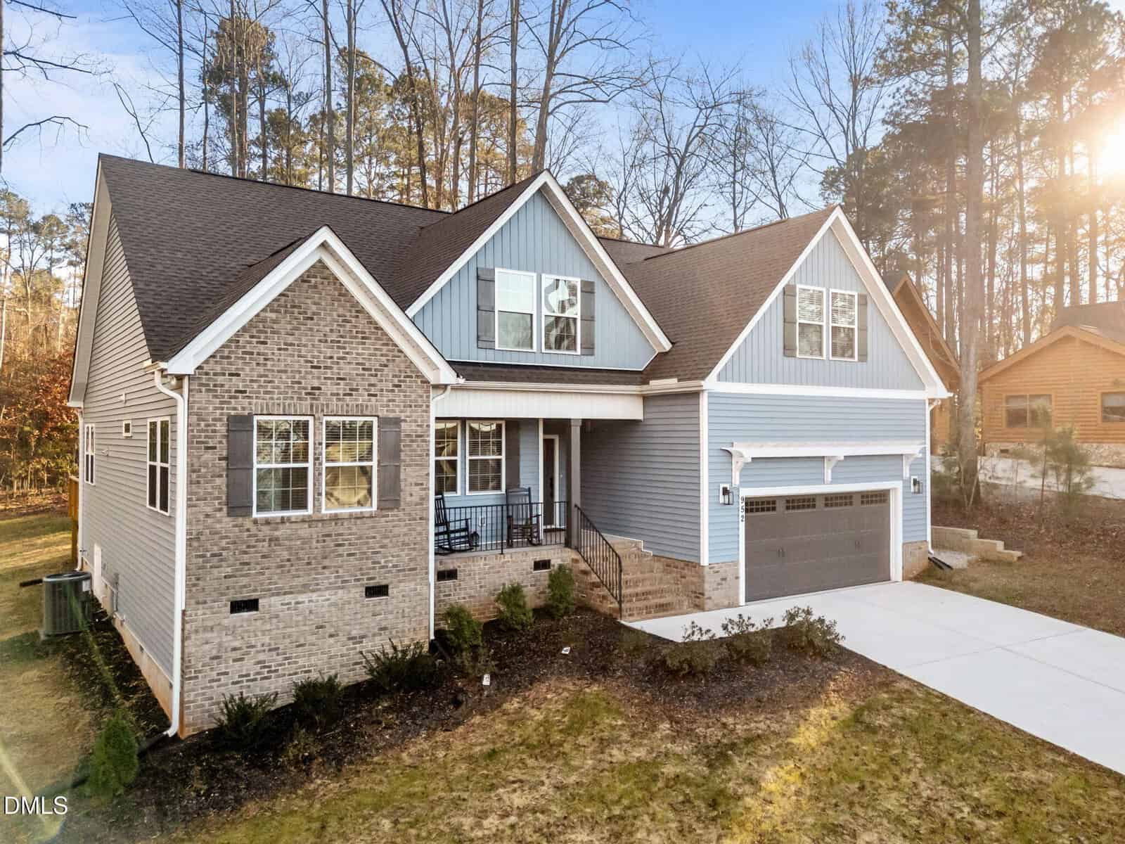 Two-story suburban house with gray brick and blue siding exterior, covered front porch, attached two-car garage, and concrete driveway. Surrounded by trees and neighboring homes under a clear sky.