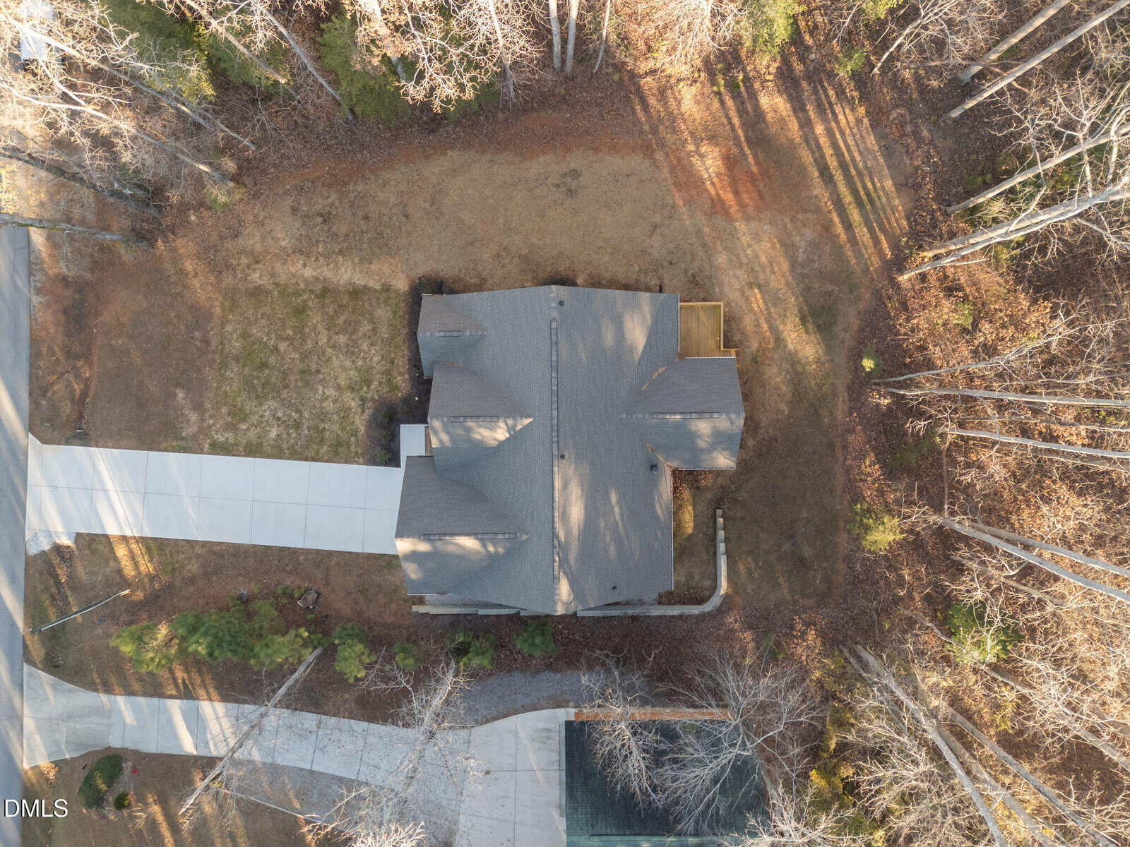 Aerial view of a single-story house with a gray roof, a driveway, and a surrounding yard bordered by trees. The property is situated in a wooded residential area with visible shadows from the trees.