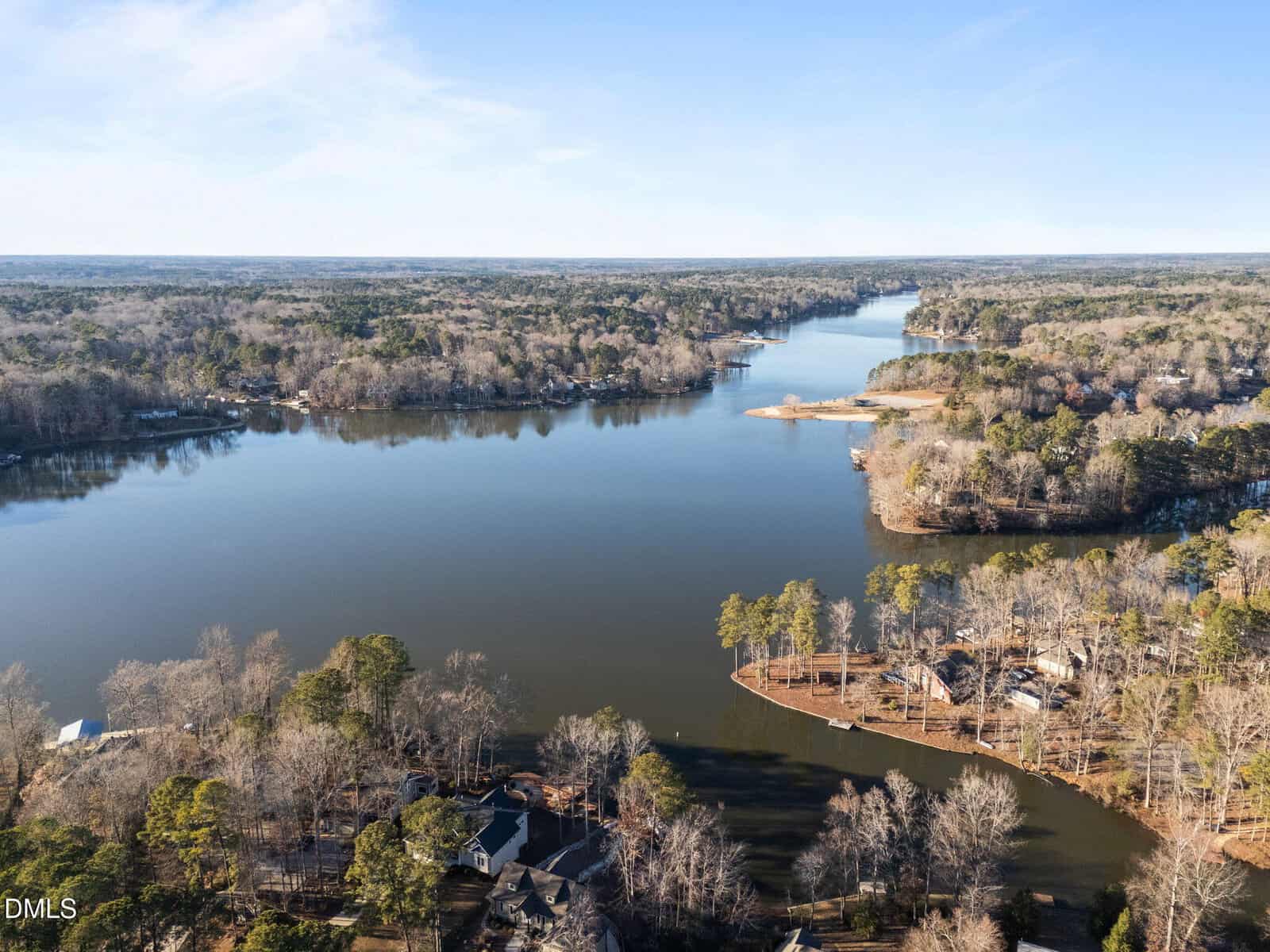 Aerial view of a wide river or lake surrounded by dense, leafless trees and several houses along the shoreline on a clear day with blue sky.