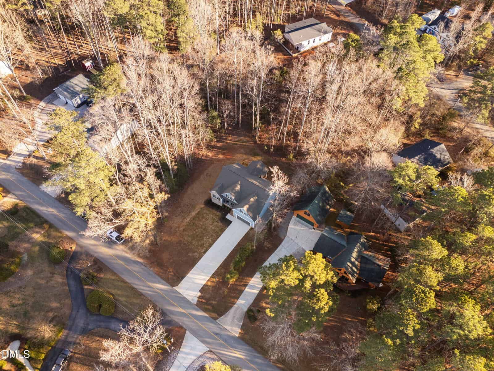 Aerial view of a suburban neighborhood with houses surrounded by trees; one house with a long driveway is centered, and neighboring houses and roads are visible among wooded areas.