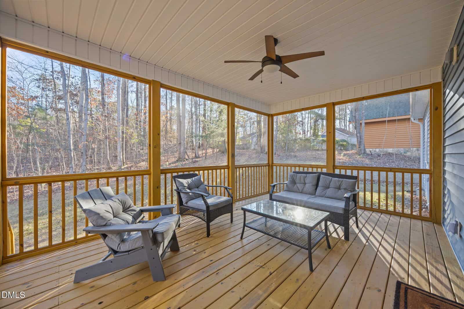 A screened-in porch with wooden floors, a ceiling fan, outdoor furniture including two cushioned chairs, a loveseat, and a glass-top coffee table, overlooking a wooded backyard.