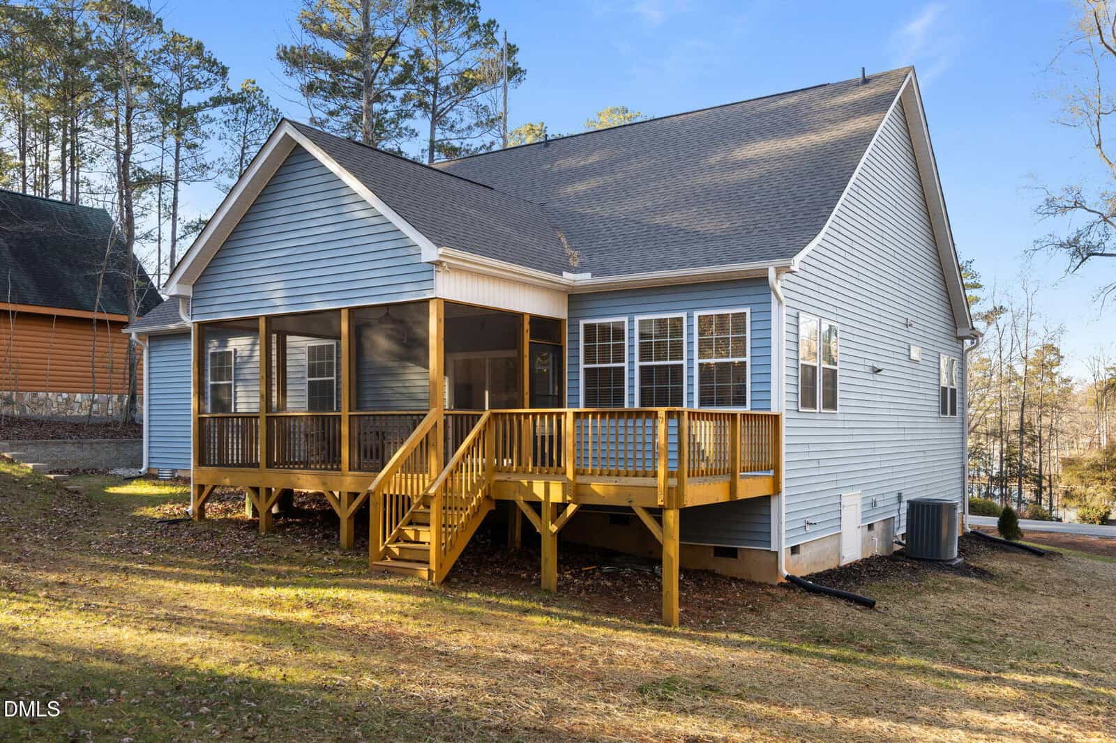A light blue single-story house with a gray shingle roof, featuring a wooden deck and screened porch in the backyard. The yard is grassy with some patches of dirt, and trees surround the property.