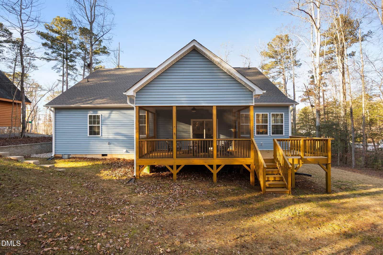 A blue house with a gray roof features a large screened-in porch and attached wooden deck with stairs, surrounded by trees and a grassy yard covered in fallen leaves.