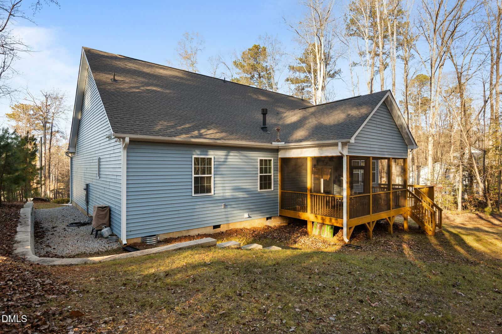 A blue house with gray shingles, a screened porch on a wooden deck, and a sloped backyard bordered by trees. The ground is grassy with some fallen leaves, and there is a gravel area along one side of the house.