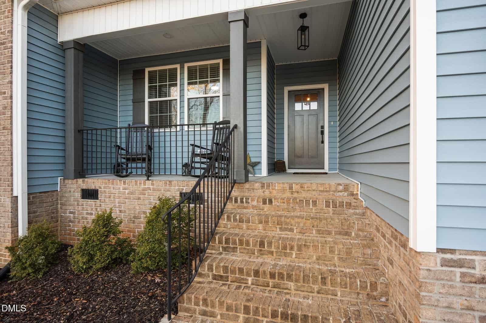 A brick staircase with a black metal railing leads to a covered porch with two black rocking chairs. The house exterior is light blue siding with white trim and a gray front door. Shrubs are planted along the foundation.