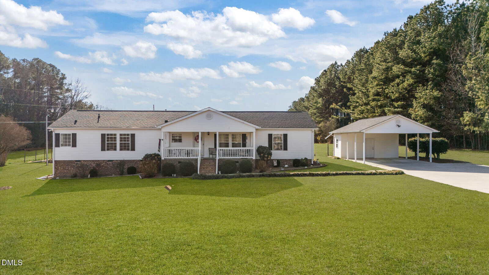 A single-story white house with a front porch and attached brick foundation sits on a large, well-maintained green lawn. A detached carport with a driveway is on the right, and trees border the property in the background.
