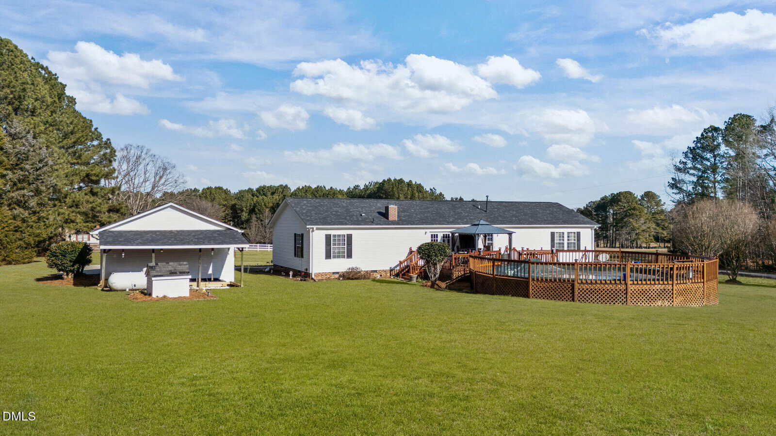 A single-story white house with a dark roof sits on a large, green lawn. There is a separate garage to the left and a round above-ground pool with a wooden deck on the right. Trees border the yard under a partly cloudy sky.