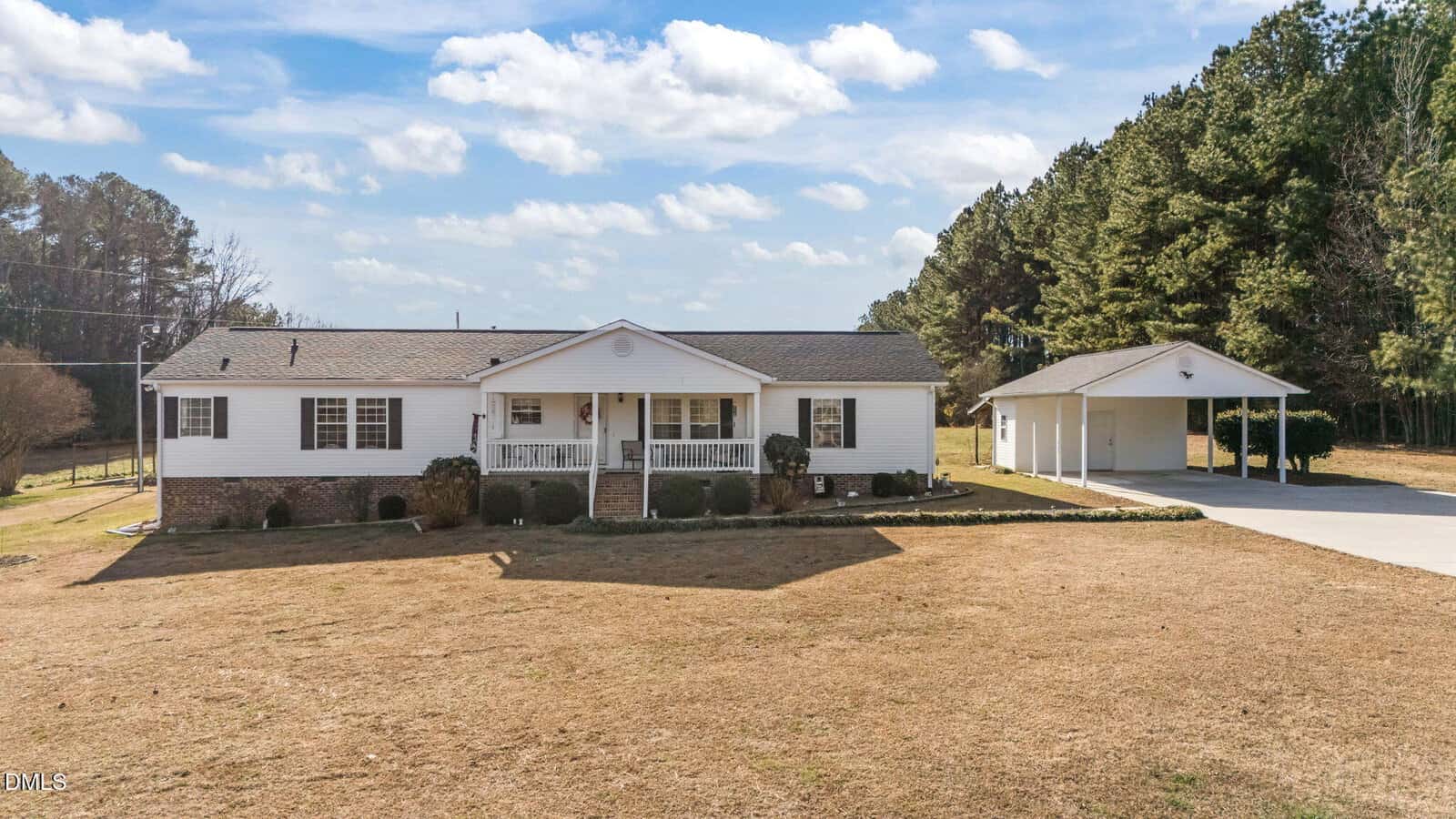 A single-story white house with a front porch and gable roof, set on a large grassy lot with minimal landscaping. A detached covered carport is on the right, bordered by tall trees in the background under a partly cloudy sky.