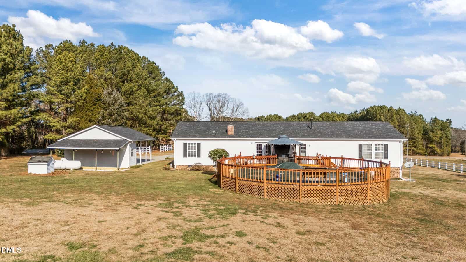 A single-story white house with a dark roof sits on a large grassy lot. There is a wooden deck surrounding an above-ground pool and a detached covered structure nearby. Pine trees and a white fence are in the background.