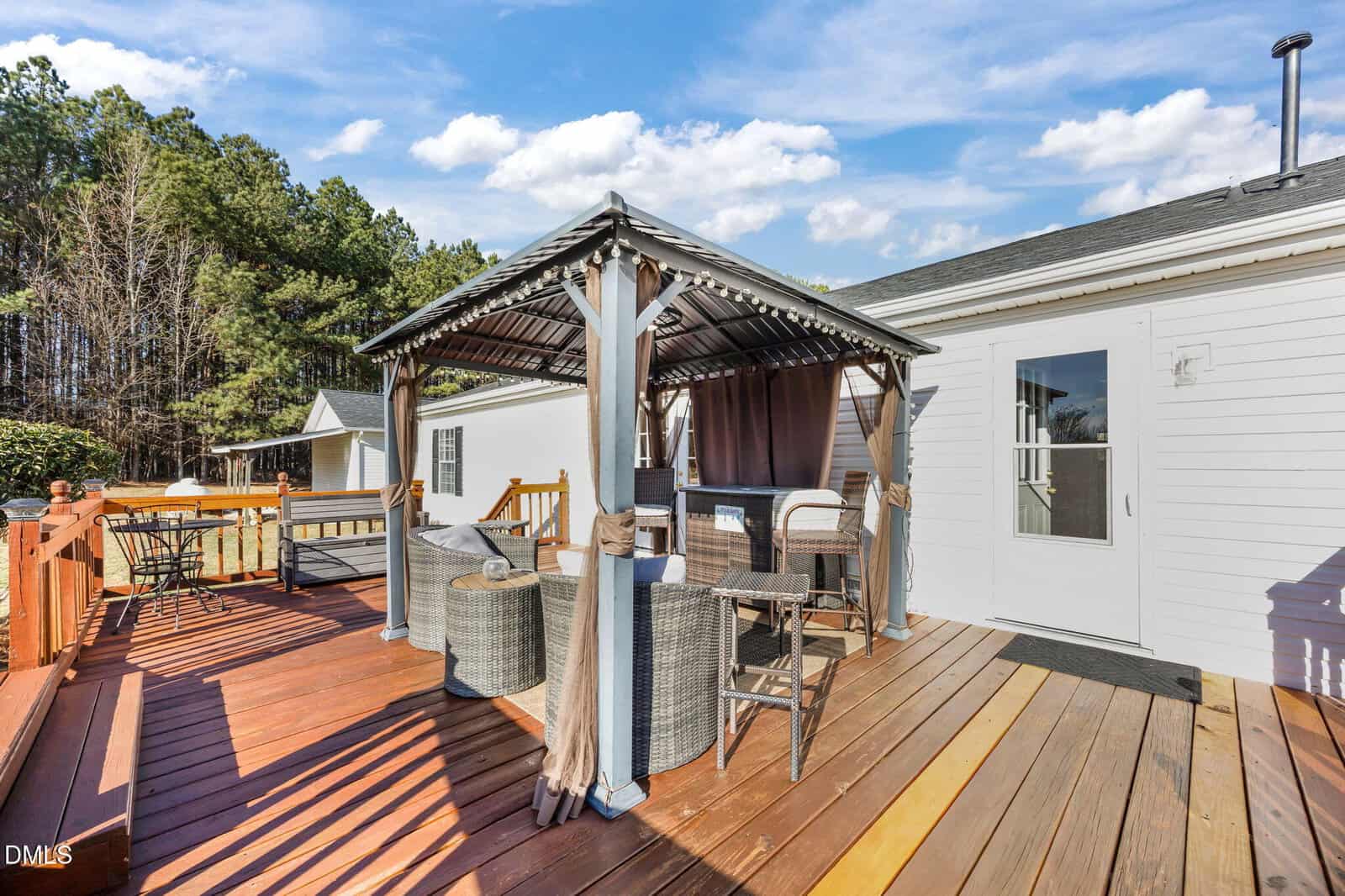 A wooden deck with outdoor seating under a gazebo, connected to a white house. The deck features a table, wicker chairs, and has trees and blue sky in the background.