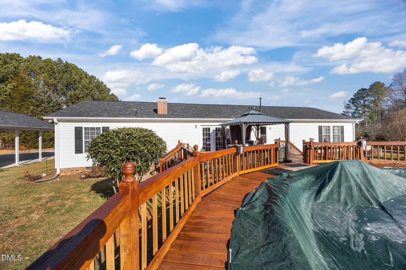 A single-story white house with a gray roof, wooden deck, and covered above-ground pool in the backyard. Trees and blue sky with clouds are in the background.