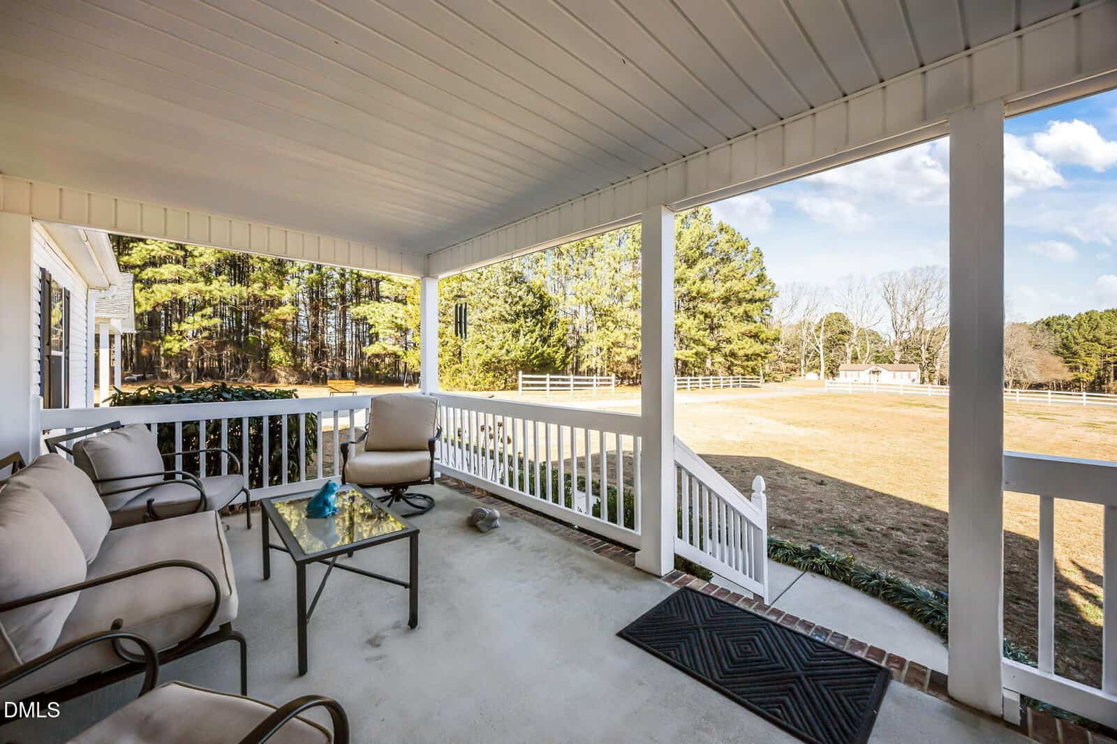 A covered porch with cushioned chairs, a glass coffee table, and a view of a spacious, grassy yard bordered by trees and white fencing on a sunny day.