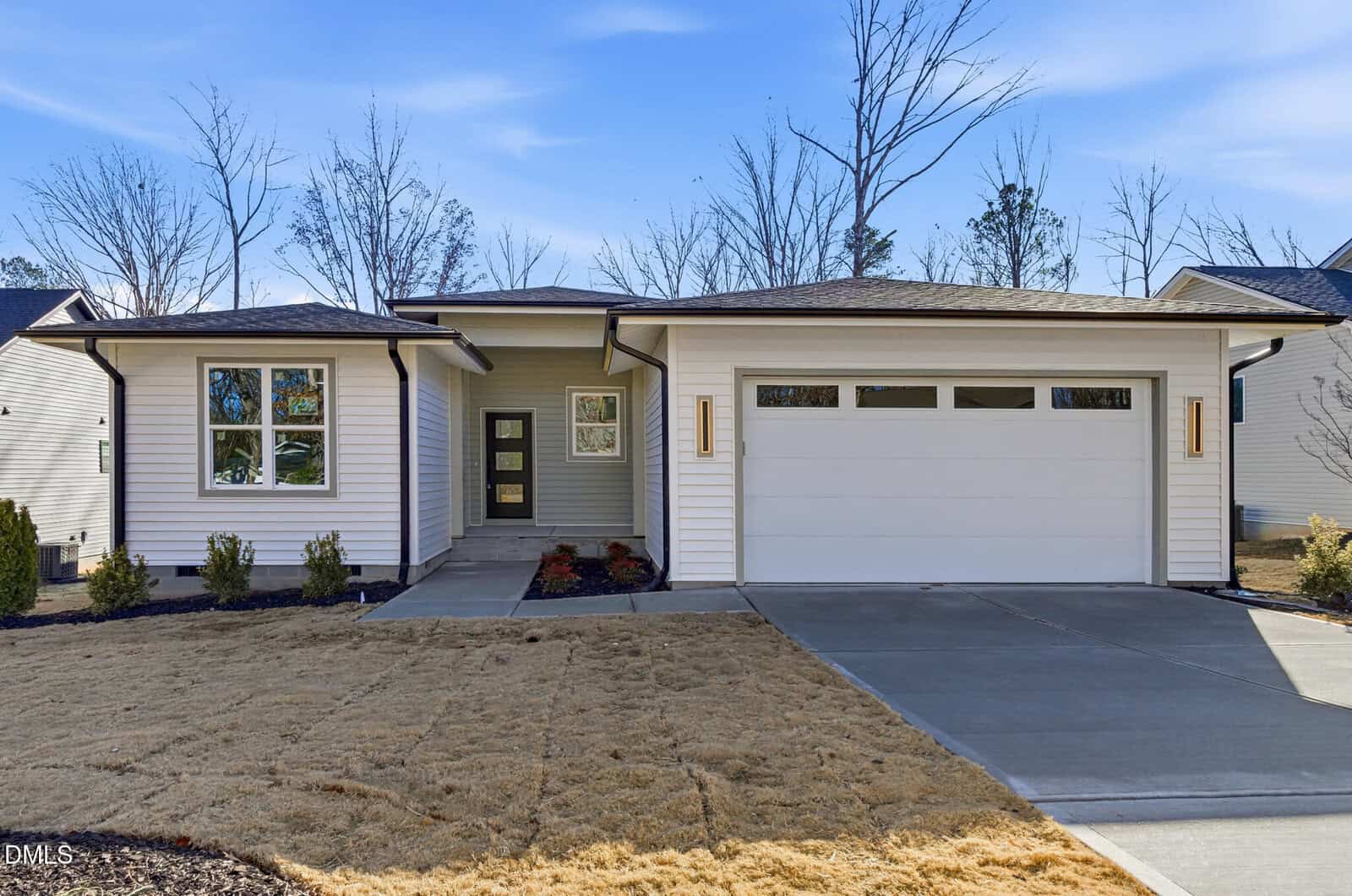 Modern single-story house at 646 Shawnee Drive, Louisburg, with white siding, a two-car garage, and a covered front entrance. The yard features minimal landscaping with brown grass and a few shrubs; leafless trees stand under a blue sky.
