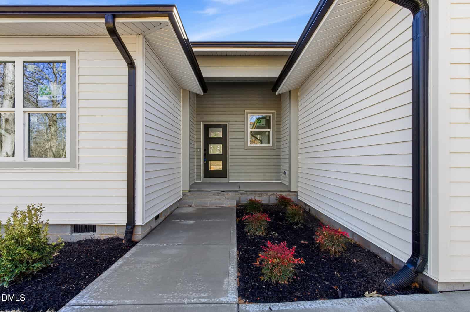 A concrete walkway leads to a modern front door with glass panels, flanked by cream-colored siding and small landscaped garden beds with shrubs on both sides.