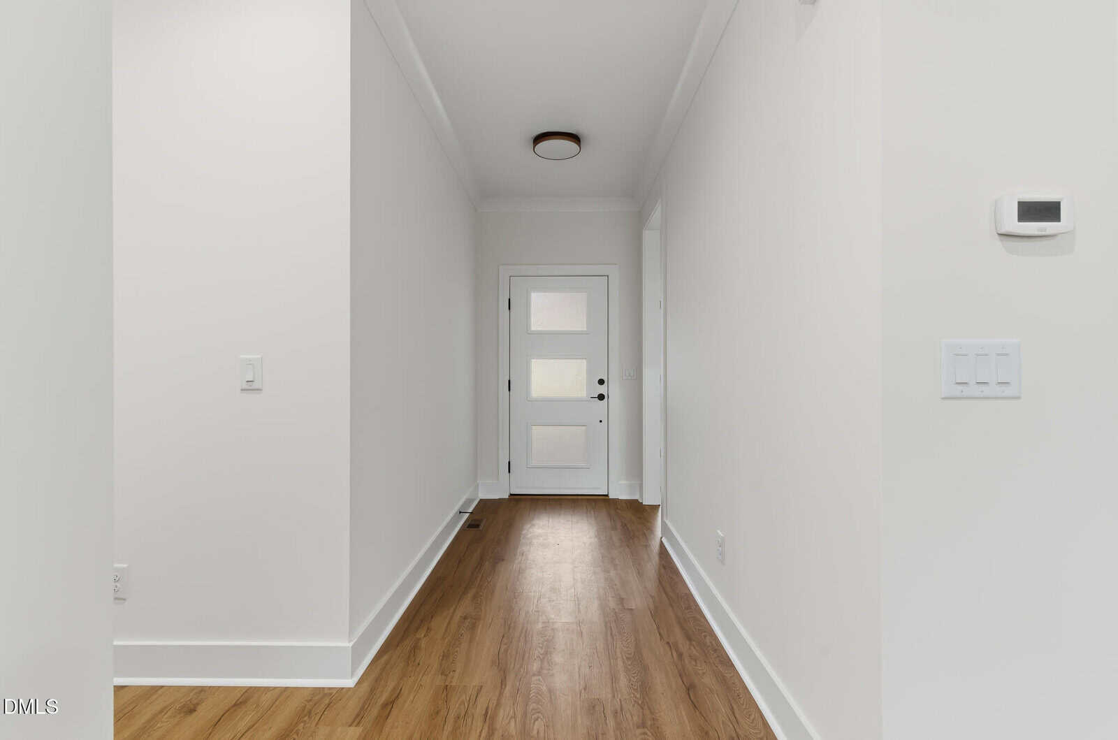 A bright, minimalist hallway with white walls and ceiling, wood flooring, a modern light fixture, and a white front door with frosted glass panels. A thermostat and light switches are visible on the right wall.