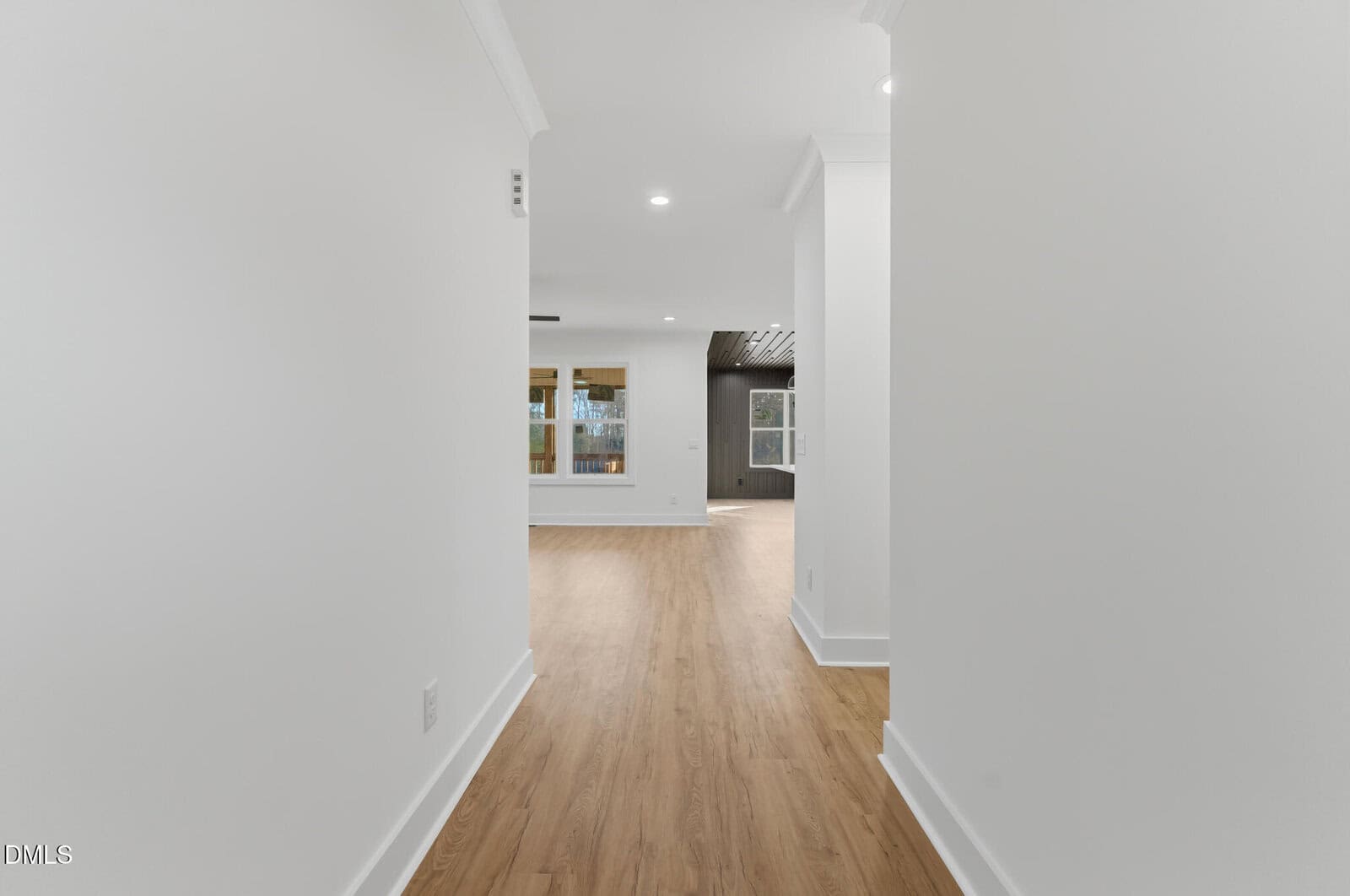 A bright hallway with white walls and light wood flooring leads to an open living area featuring large windows and additional rooms visible in the background.