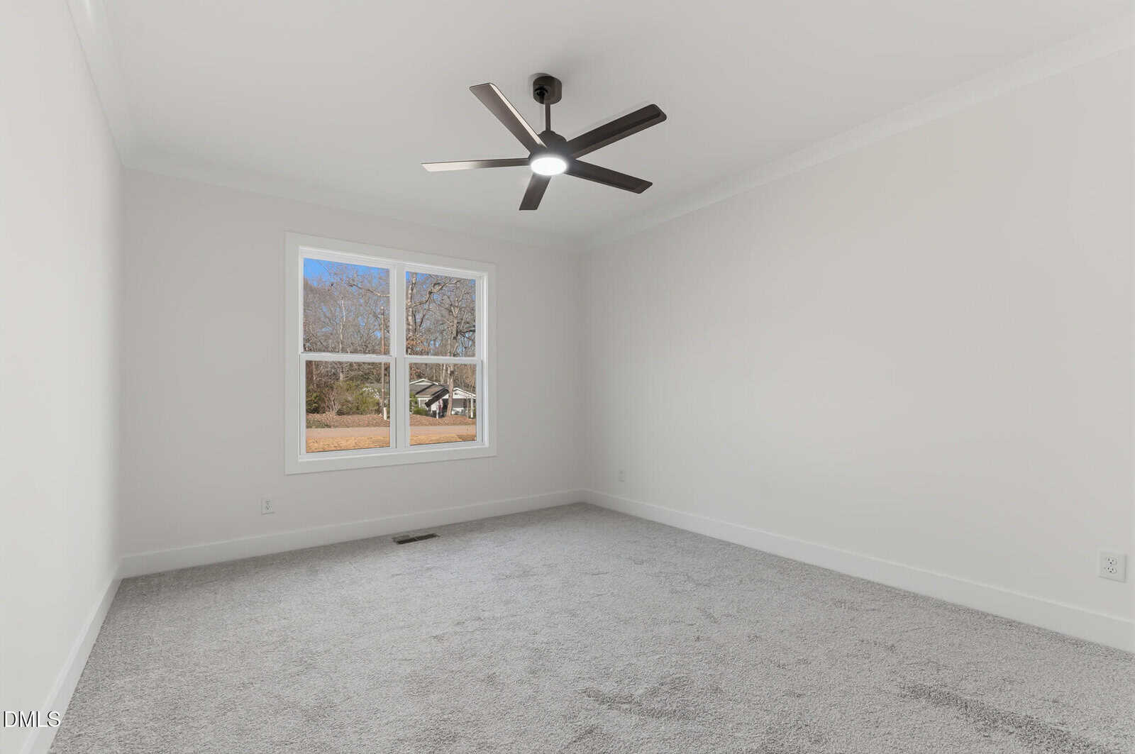 A bright, empty room with light gray carpet, white walls, a large window showing trees outside, and a black modern ceiling fan with a light.