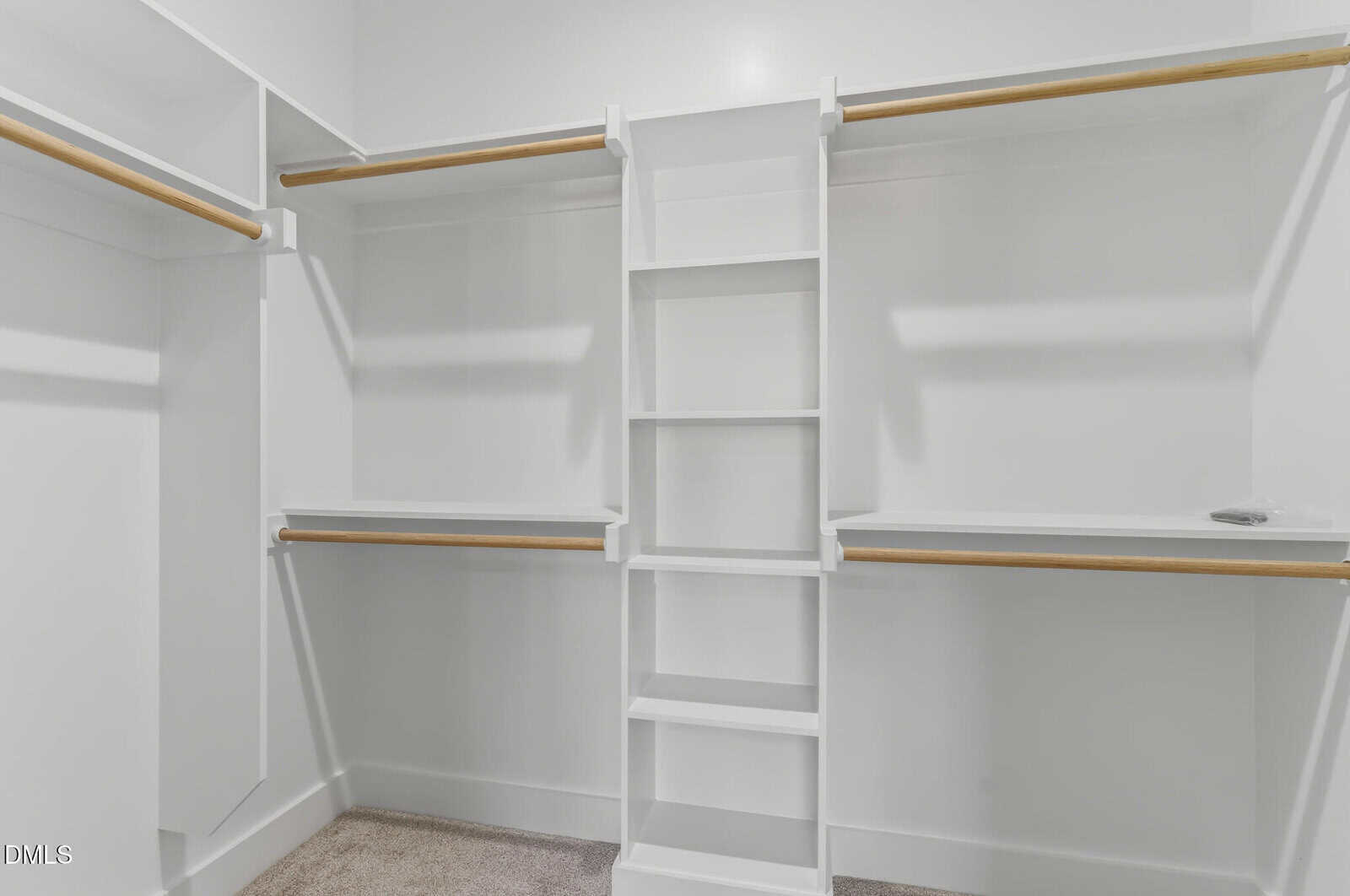 Empty walk-in closet with white walls, built-in white shelving unit in the center, wooden hanging rods on both sides, and beige carpeted floor.