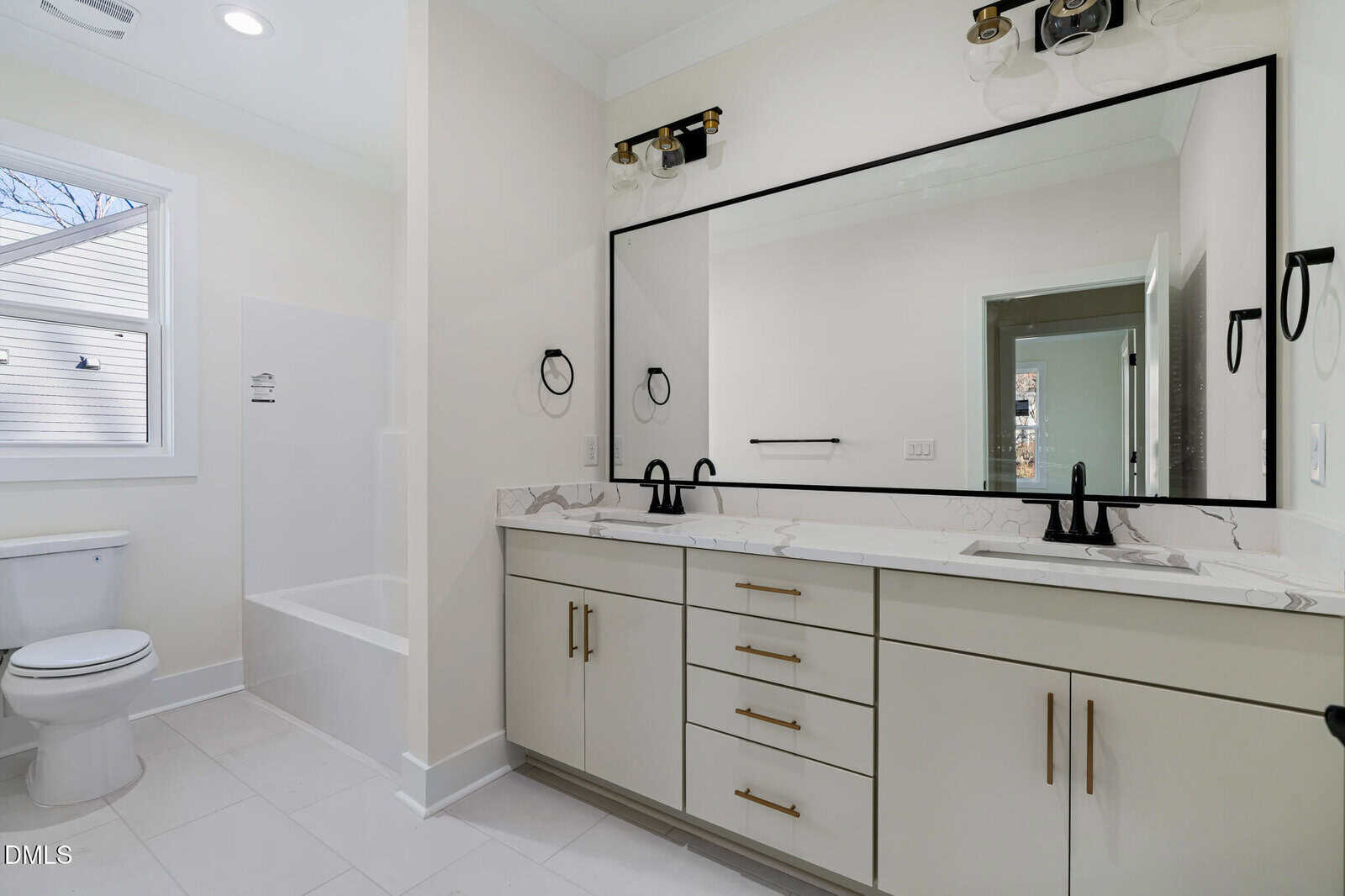 Modern bathroom with white cabinets, double sinks, black fixtures, a large mirror, a toilet, and a bathtub with a window above. The space is bright with white tile flooring and recessed lighting.