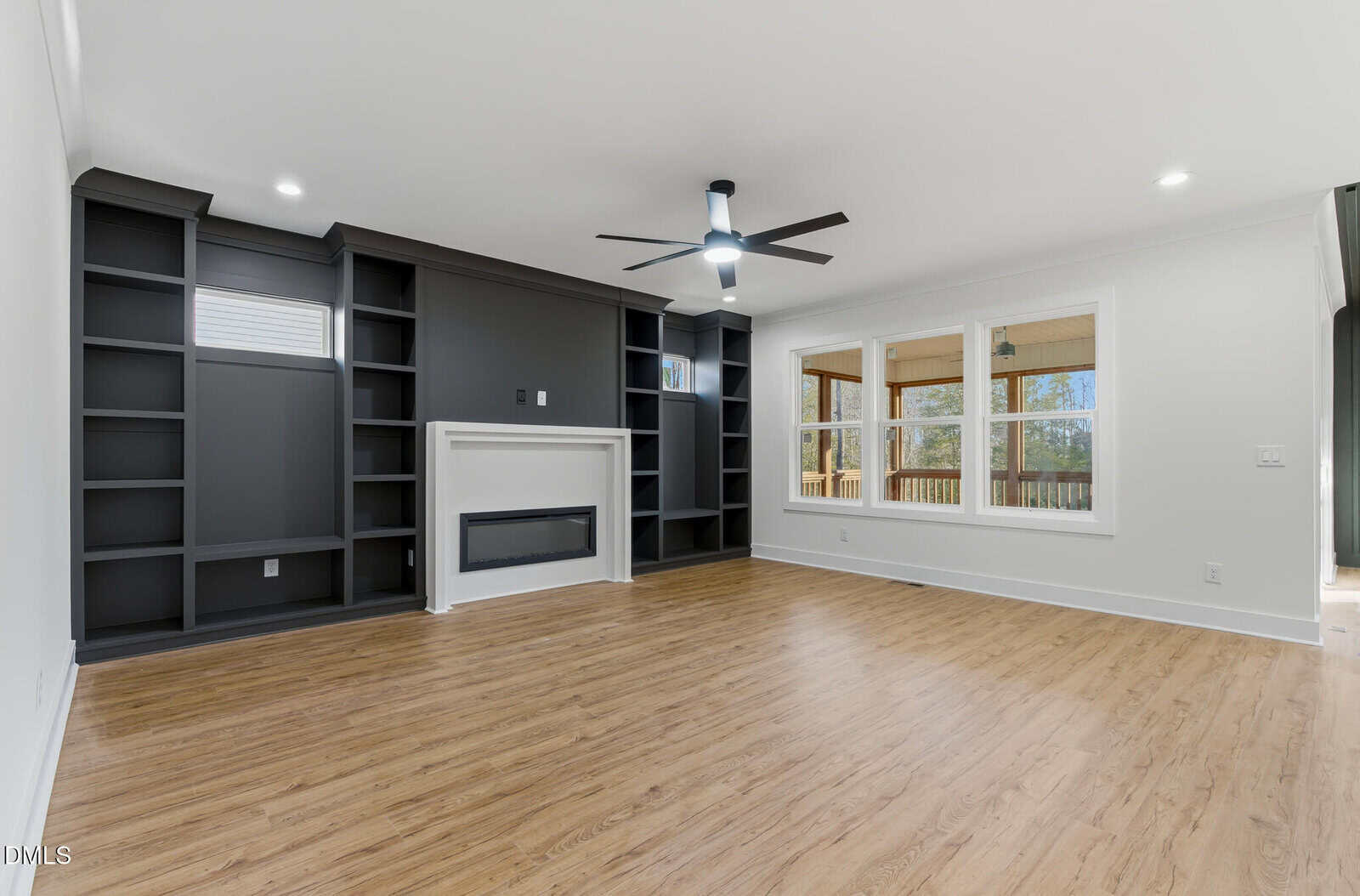A modern living room with light wood floors, dark built-in shelves flanking a fireplace, a ceiling fan, white walls, and large windows overlooking a covered porch.
